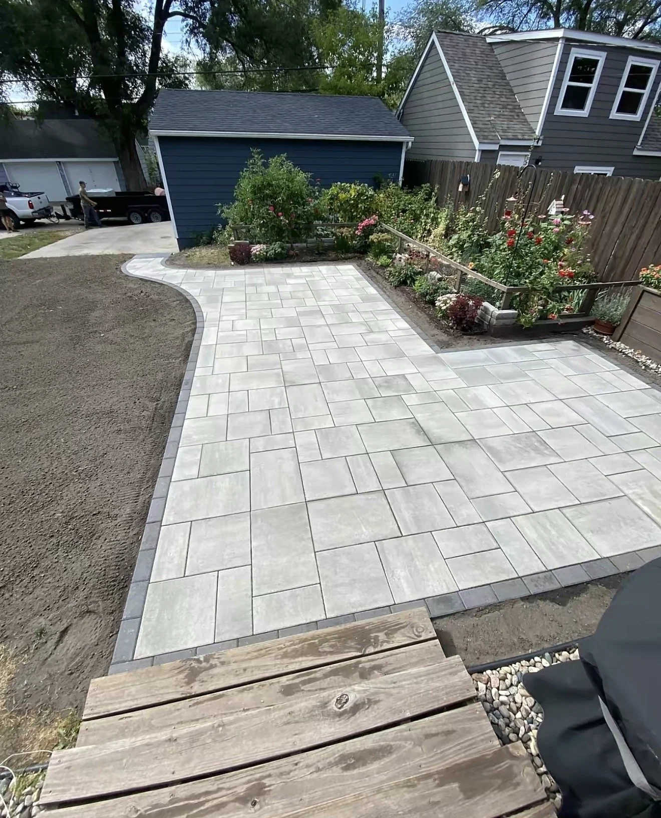 A backyard with a newly-paved walkway and garden bed filled with flowers beside a wooden fence. There are houses and parked vehicles in the background.