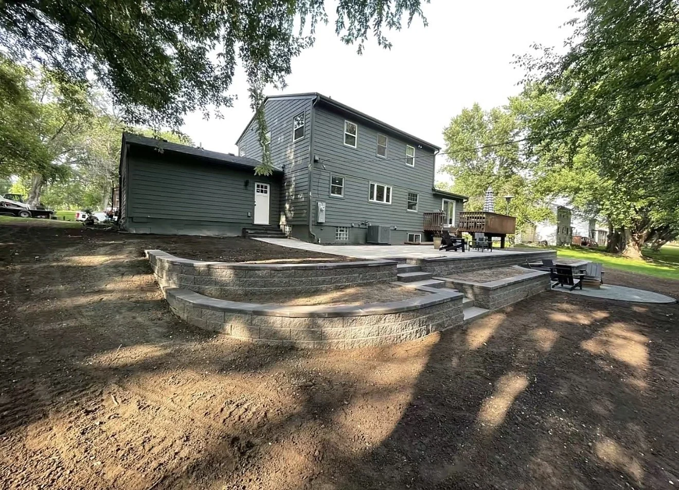 Backyard with tiered stone patio and outdoor seating area behind a gray house, shaded by large trees, with a lawn and neighboring houses in the background.