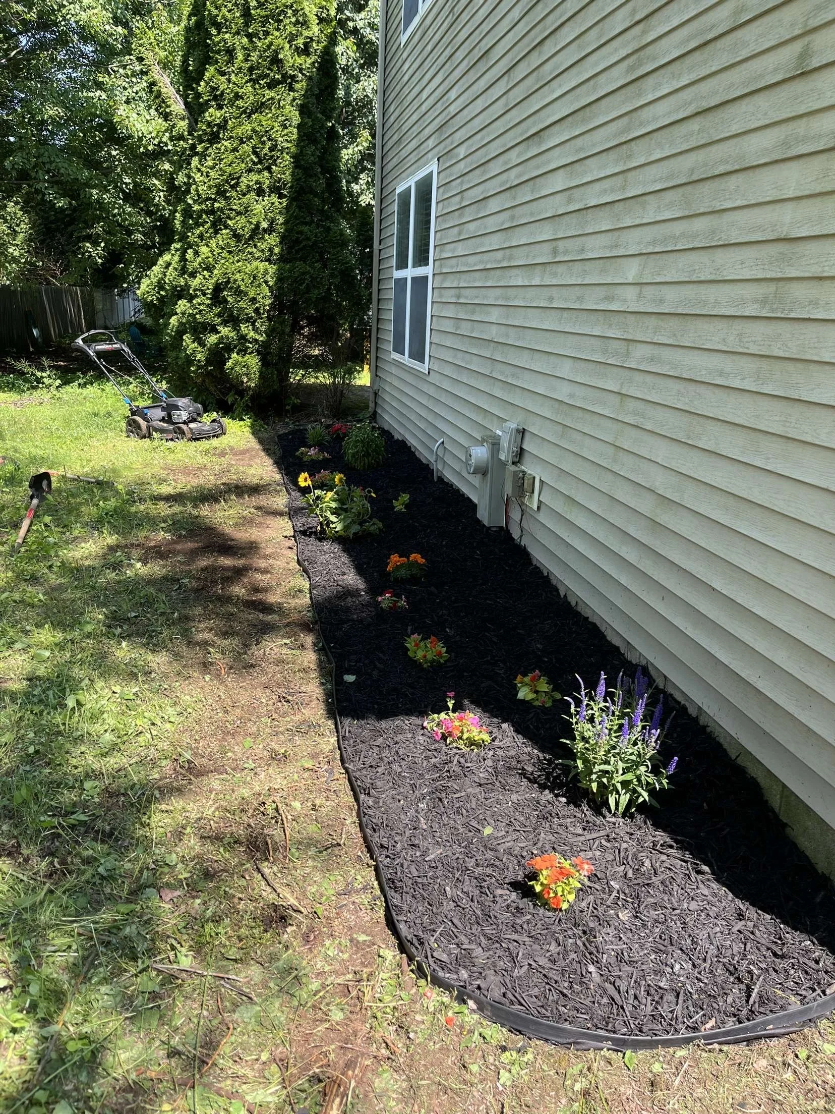 A backyard garden with a flower bed next to a beige house, featuring various colorful flowers and mulch, with a lawnmower and gardening tools nearby.