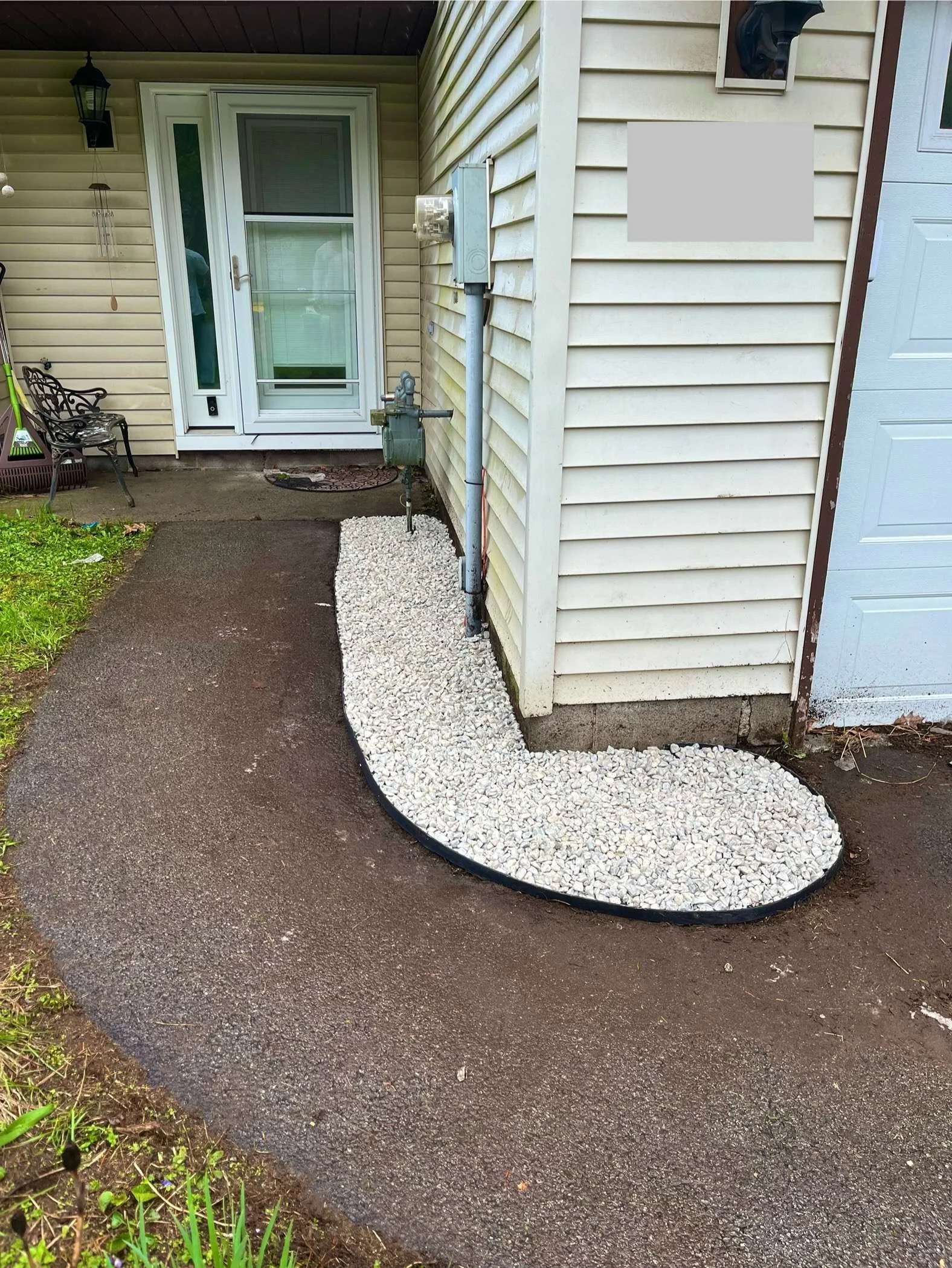 A front porch area with a driveway, a small gravel garden bed along the house's siding, a bench and a potted plant to the left, and the house's entrance with a sliding glass door and a screen door.