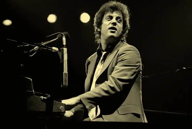 Black and white photo of a man playing the piano on stage with a microphone in front of him, under stage lights.