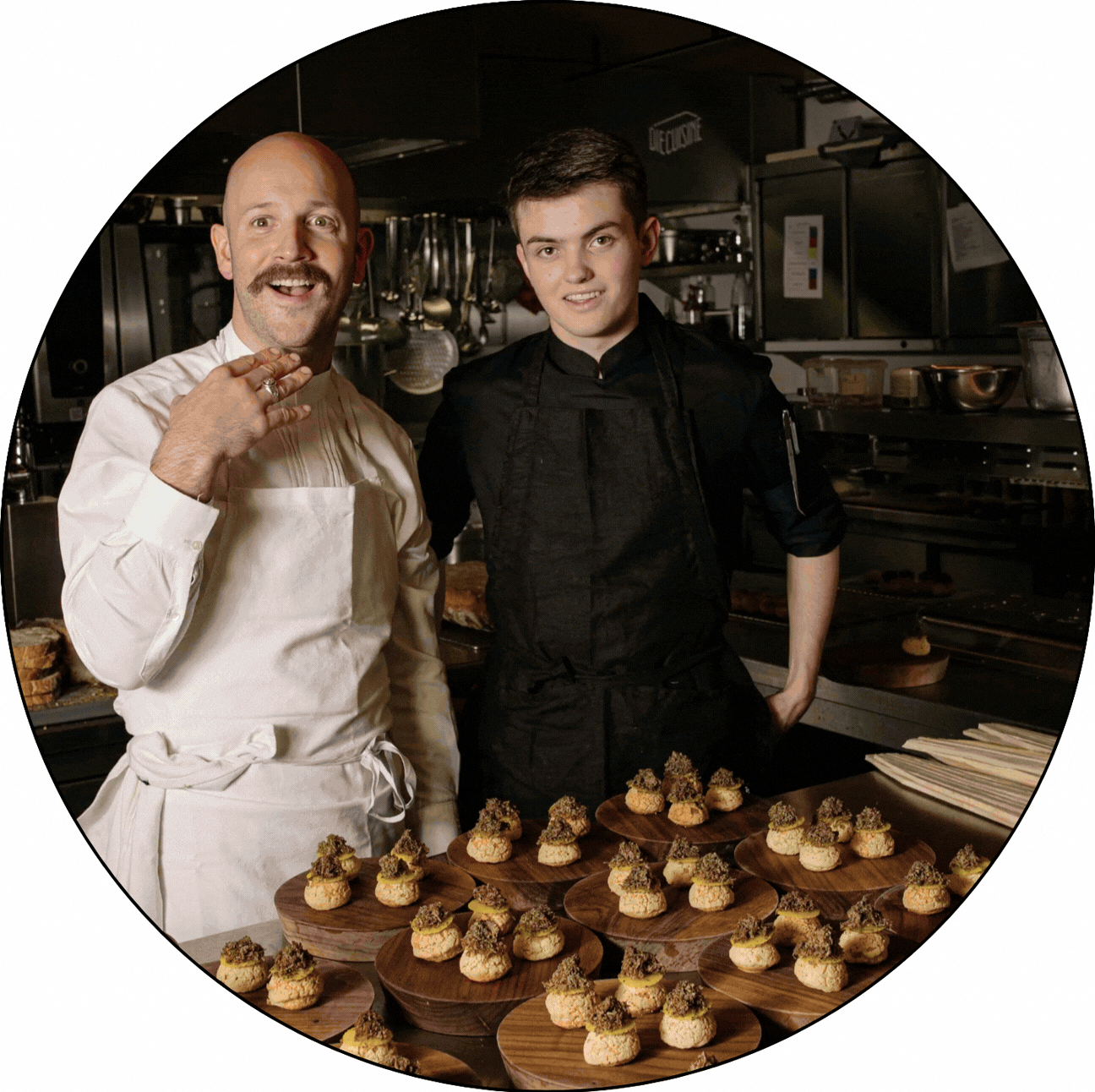 Two chefs, one older man with a bald head and a mustache, and a younger man with dark hair, standing in a professional kitchen behind a table of plated small desserts topped with garnishes.
