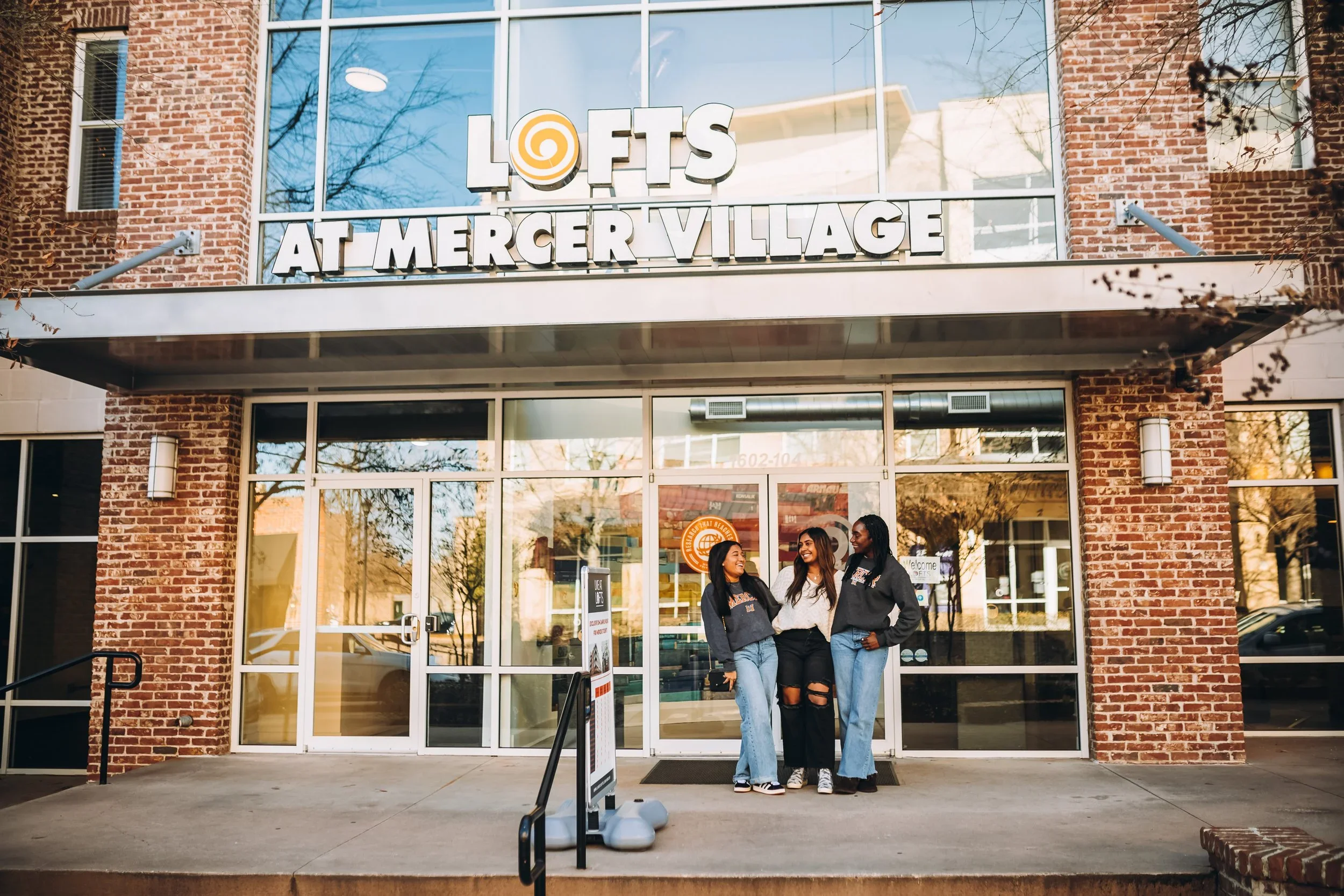 Students outside of Lofts at Mercer Village smiling in front of glass doors leading into the community area of the apartment building behind them in Macon, GA.