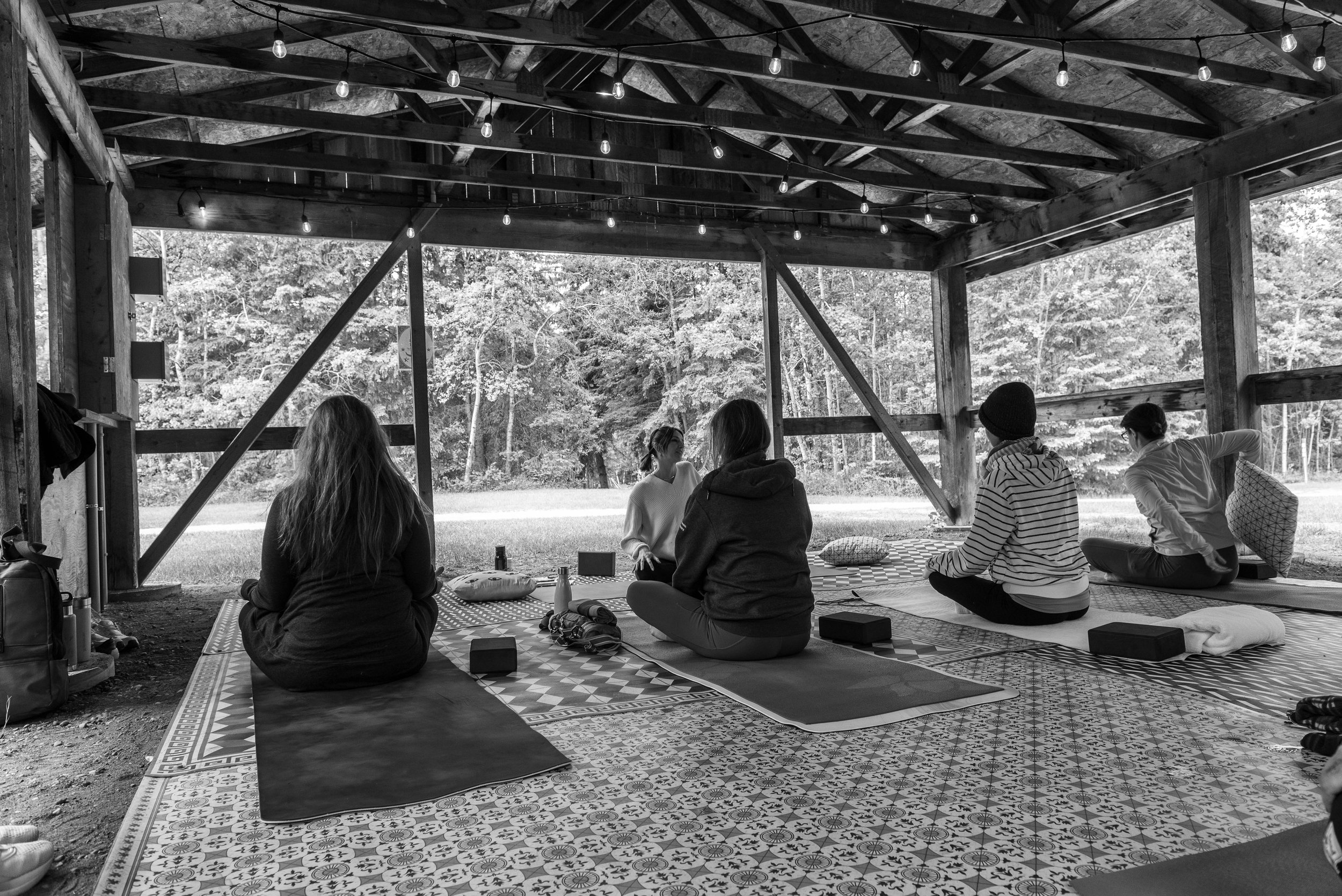 A group of woman sit on yoga mats in a comfortable seated position under a wooden shelter, surrounded by forest. Photo by Angie Leroux