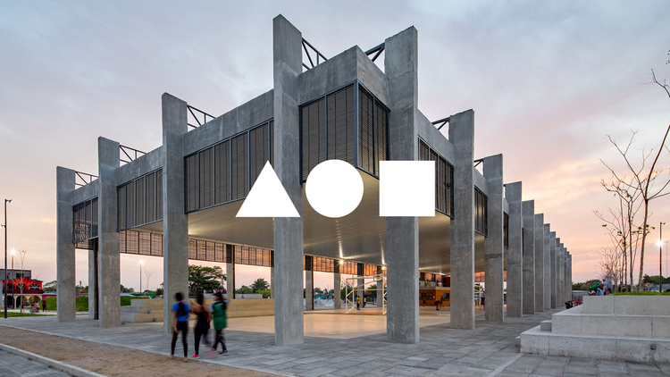 Edificio moderno de concreto con columnas grandes y ventanas de malla, bajo un cielo nublado al atardecer. Hay tres personas caminando cerca de la estructura.