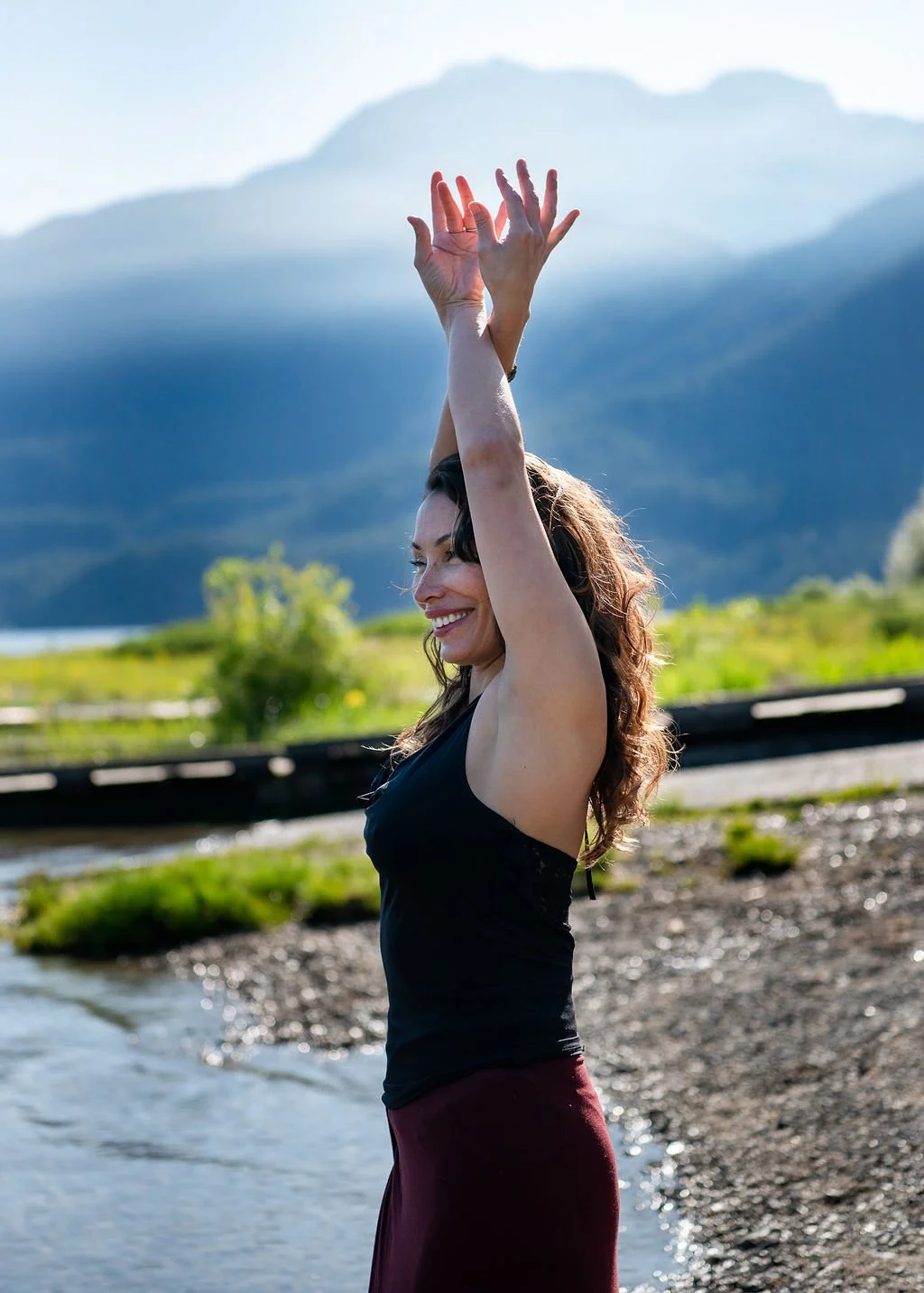 A woman standing by a river with her arms raised, smiling, with mountains and greenery in the background.