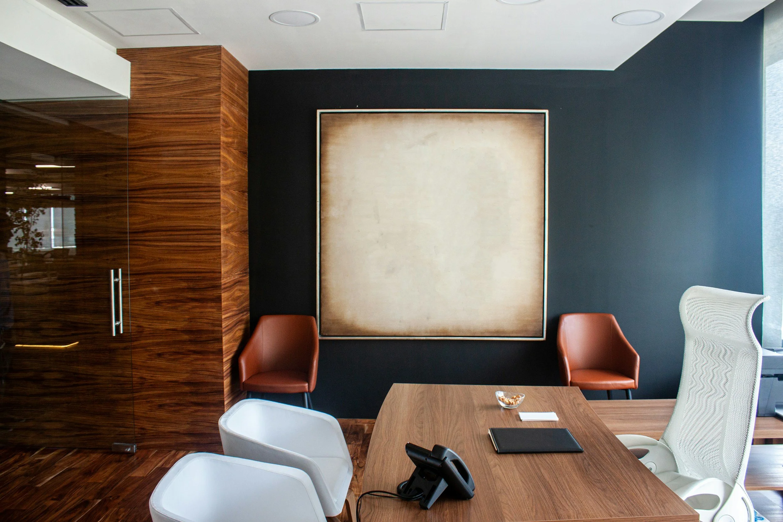 Modern office conference room with a large wooden table, white and brown chairs, a large blank artwork on a dark wall, and glass walls with wood paneling.