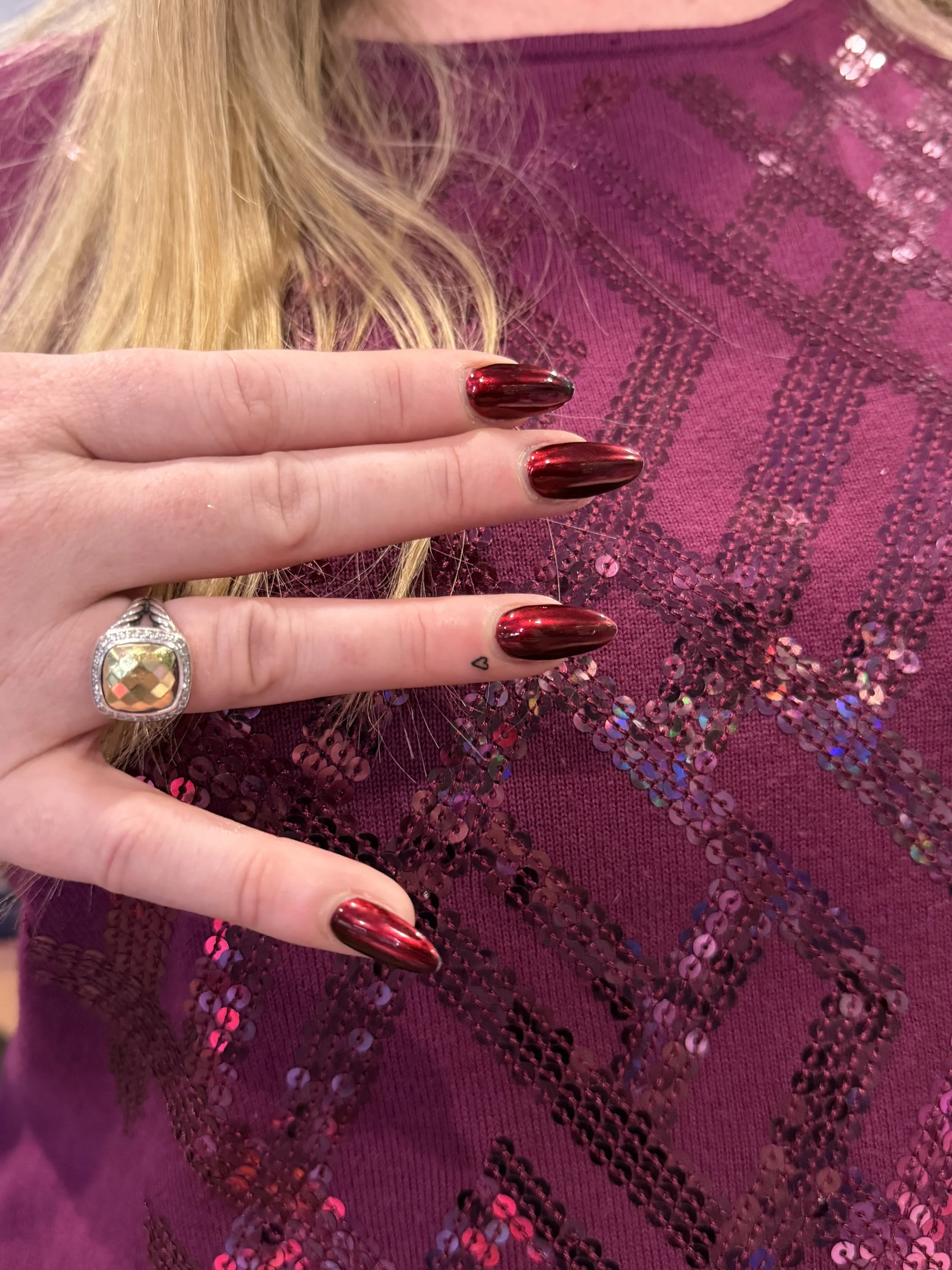 Close-up of a hand with long, dark red painted nails, wearing a silver ring with a large, faceted, yellow gemstone and a small heart tattoo on the index finger, holding a maroon fabric with shiny, sequined geometric patterns.