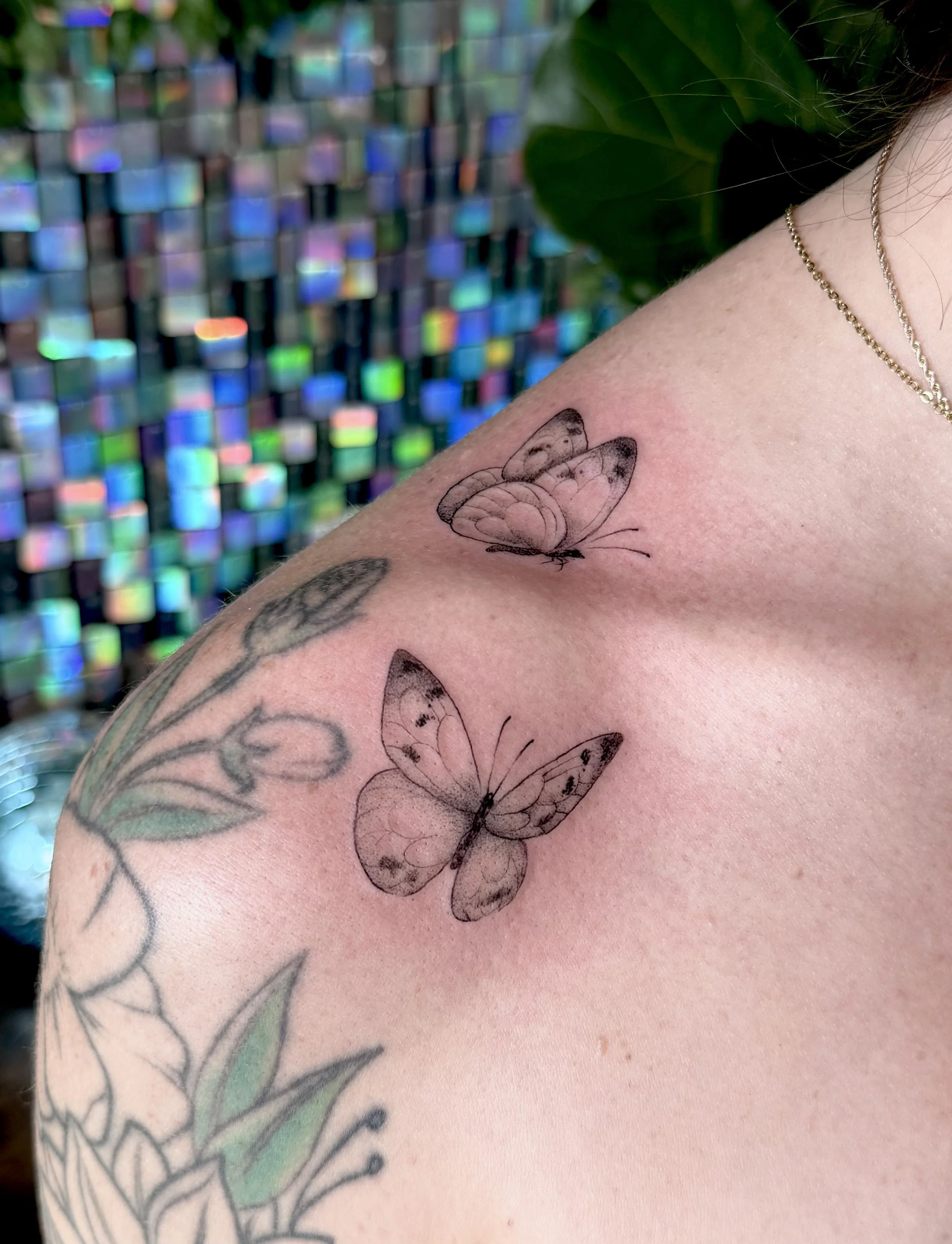 Tattoo of two butterflies on a person's shoulder, with additional tattooed flowers and leaves. The background features a holographic, iridescent pattern.