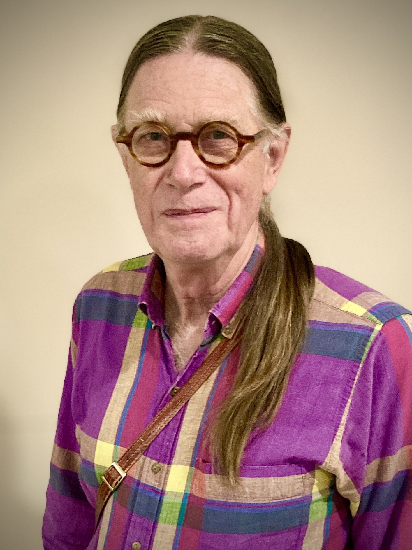 Man in a colorful shirt, glasses, and a ponytail posing against a light background.