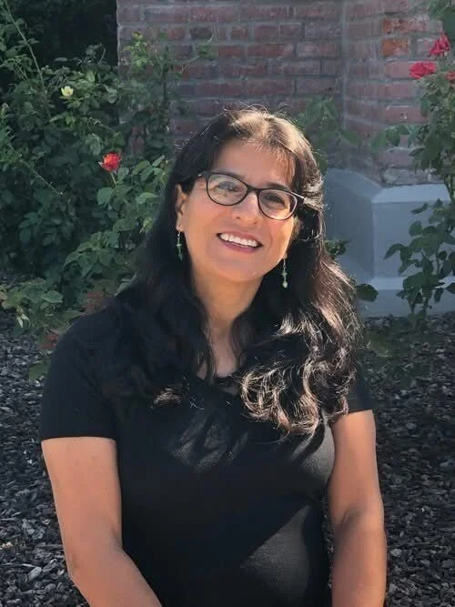 A woman with glasses and dark hair and a dark shirt, smiling, sitting in front of roses.
