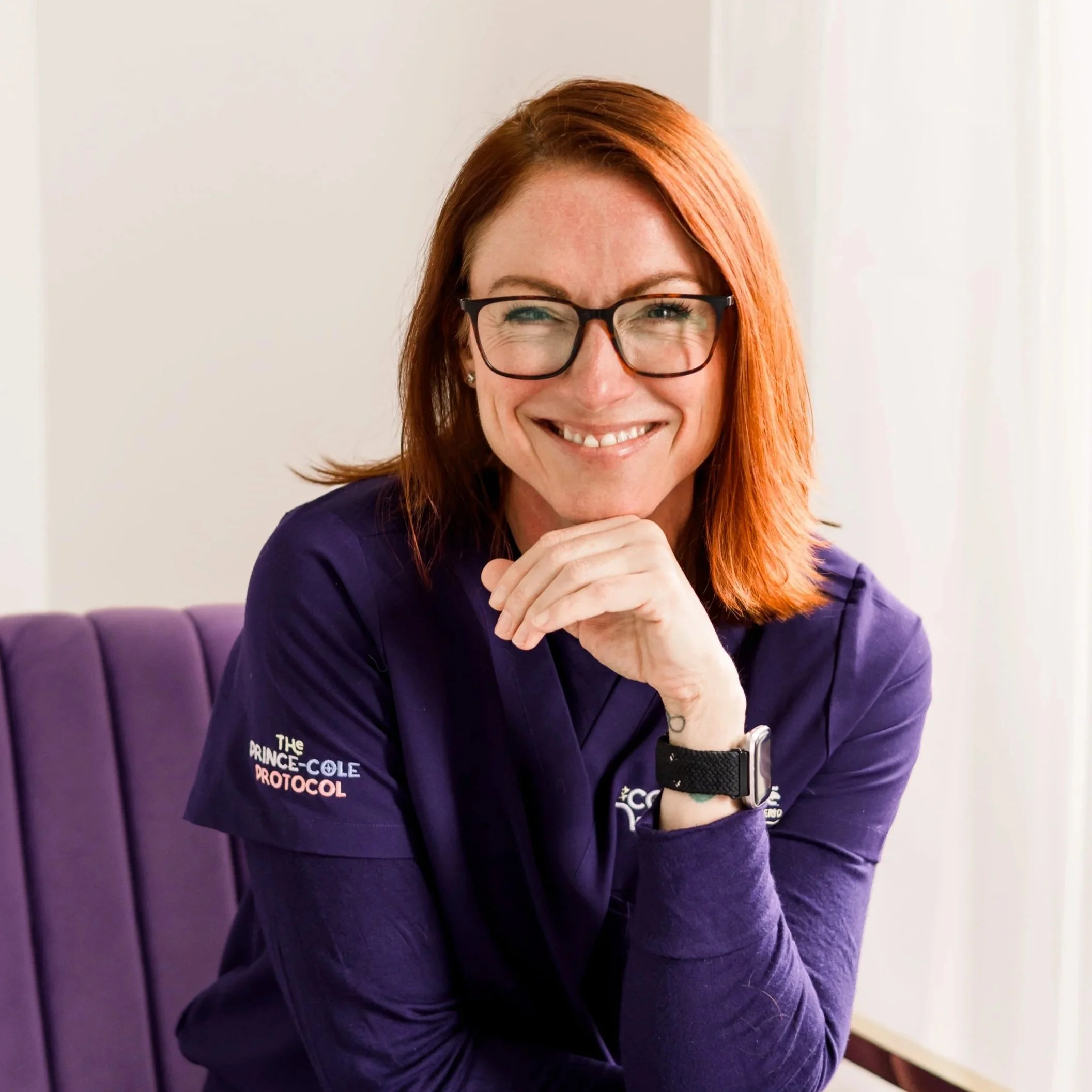 A woman with red hair, glasses, and a beige sweater, smiling and resting her chin on her hand.