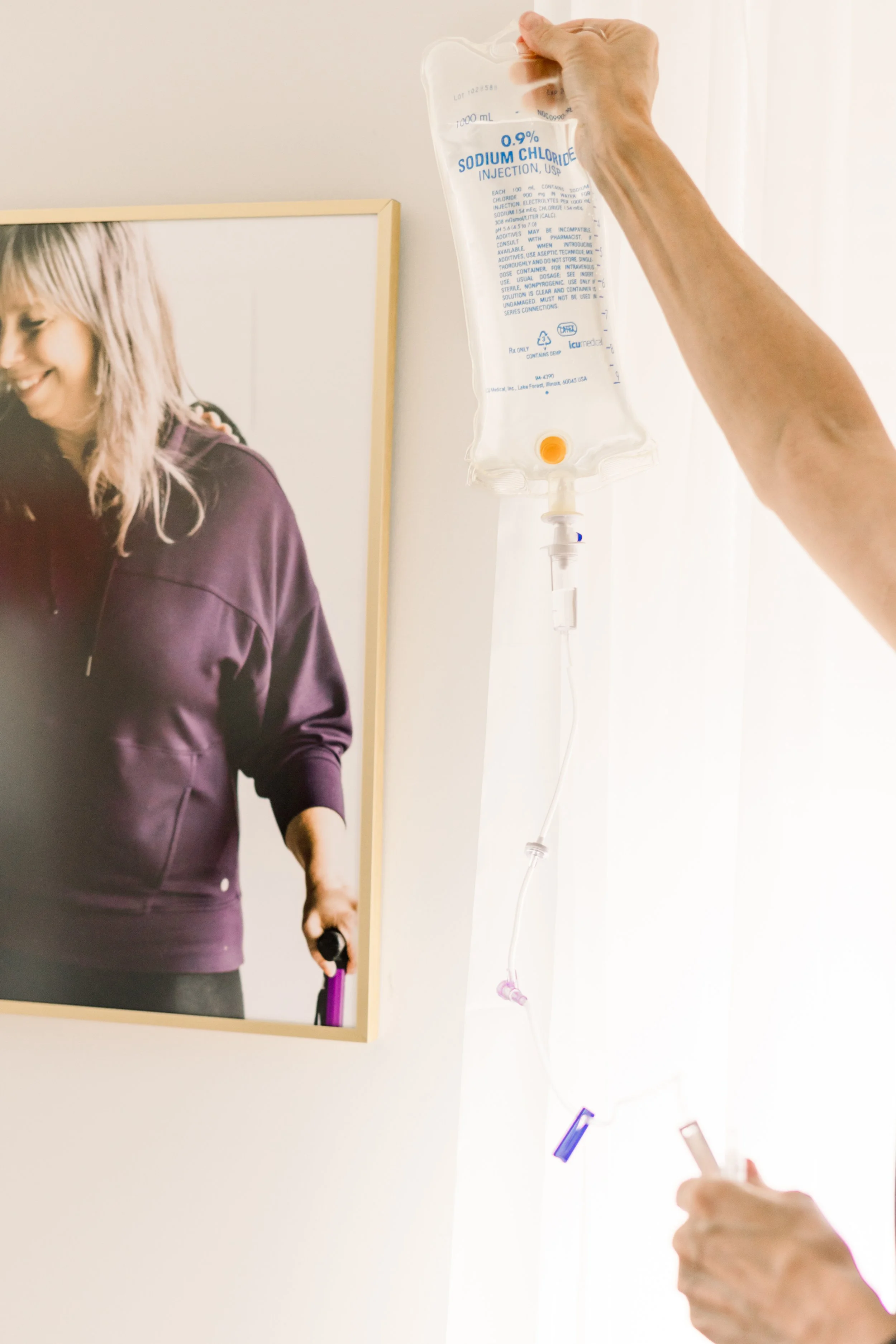 A person is hanging an intravenous (IV) fluid bag on a wall, with a framed photo of a smiling woman in a purple hoodie in the background.