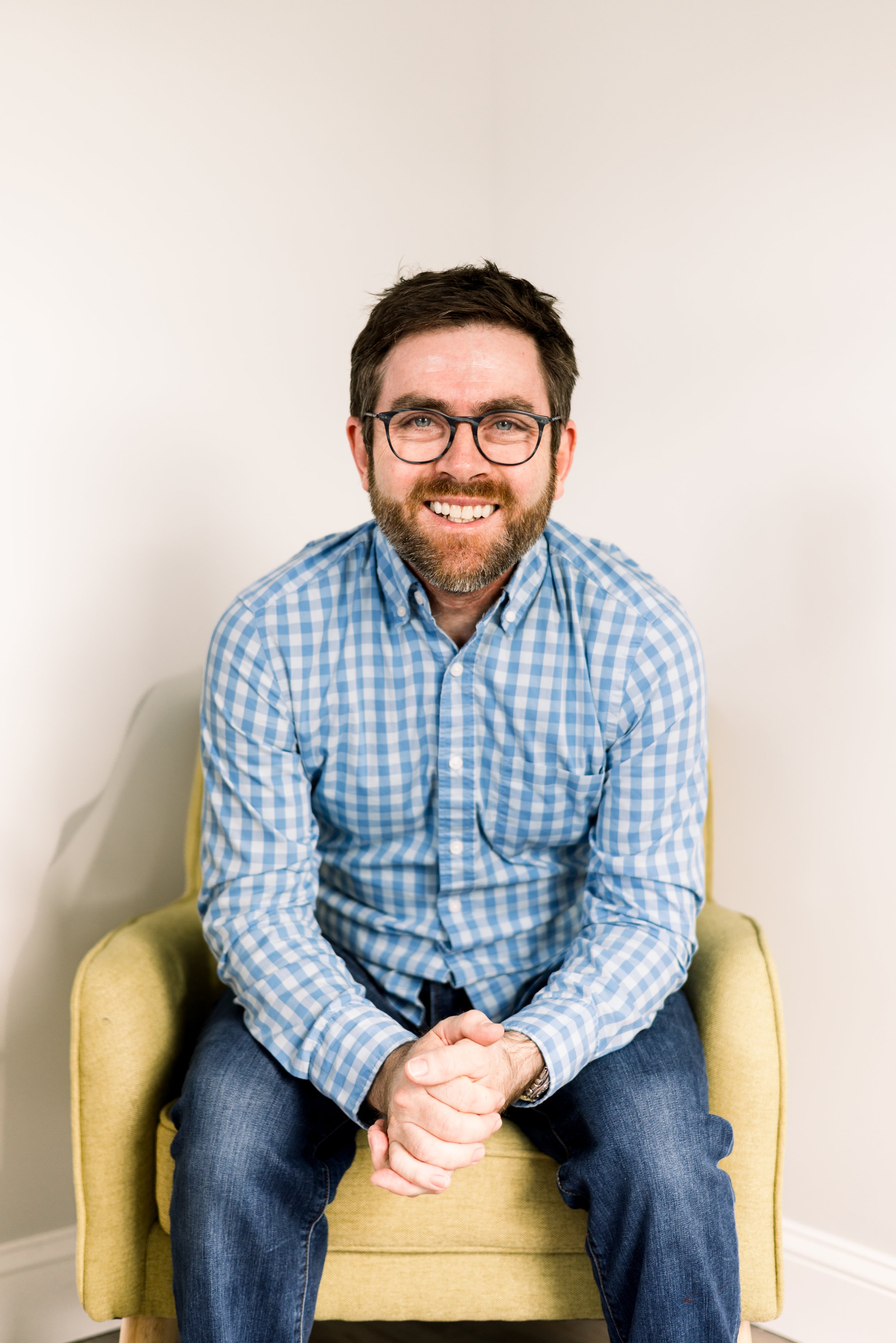 A man with glasses, a beard, and short brown hair sitting on a yellow chair against a plain white wall, smiling at the camera, wearing a blue and white checkered shirt and dark jeans.