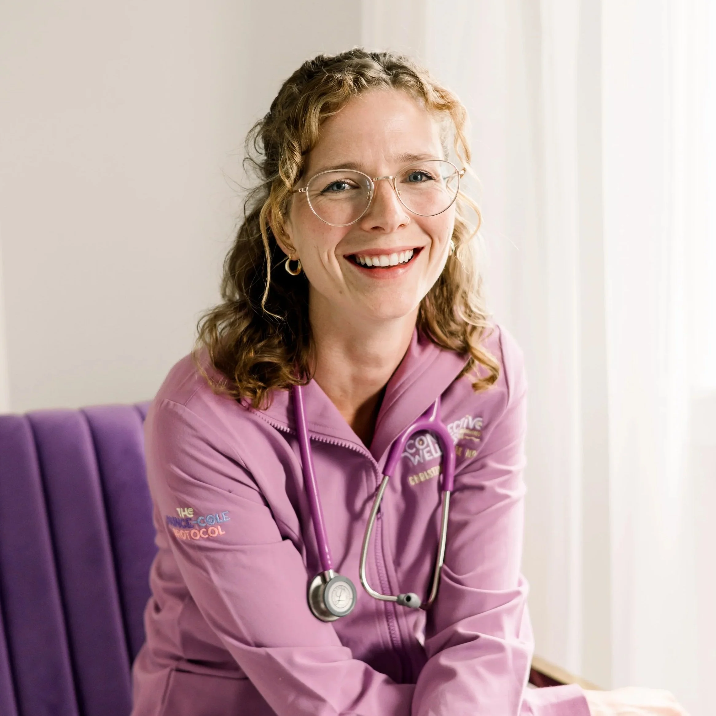 A woman with shoulder-length blonde hair, wearing glasses, a purple top, layered necklaces, and a gray and black striped cardigan, smiling in front of a white background.