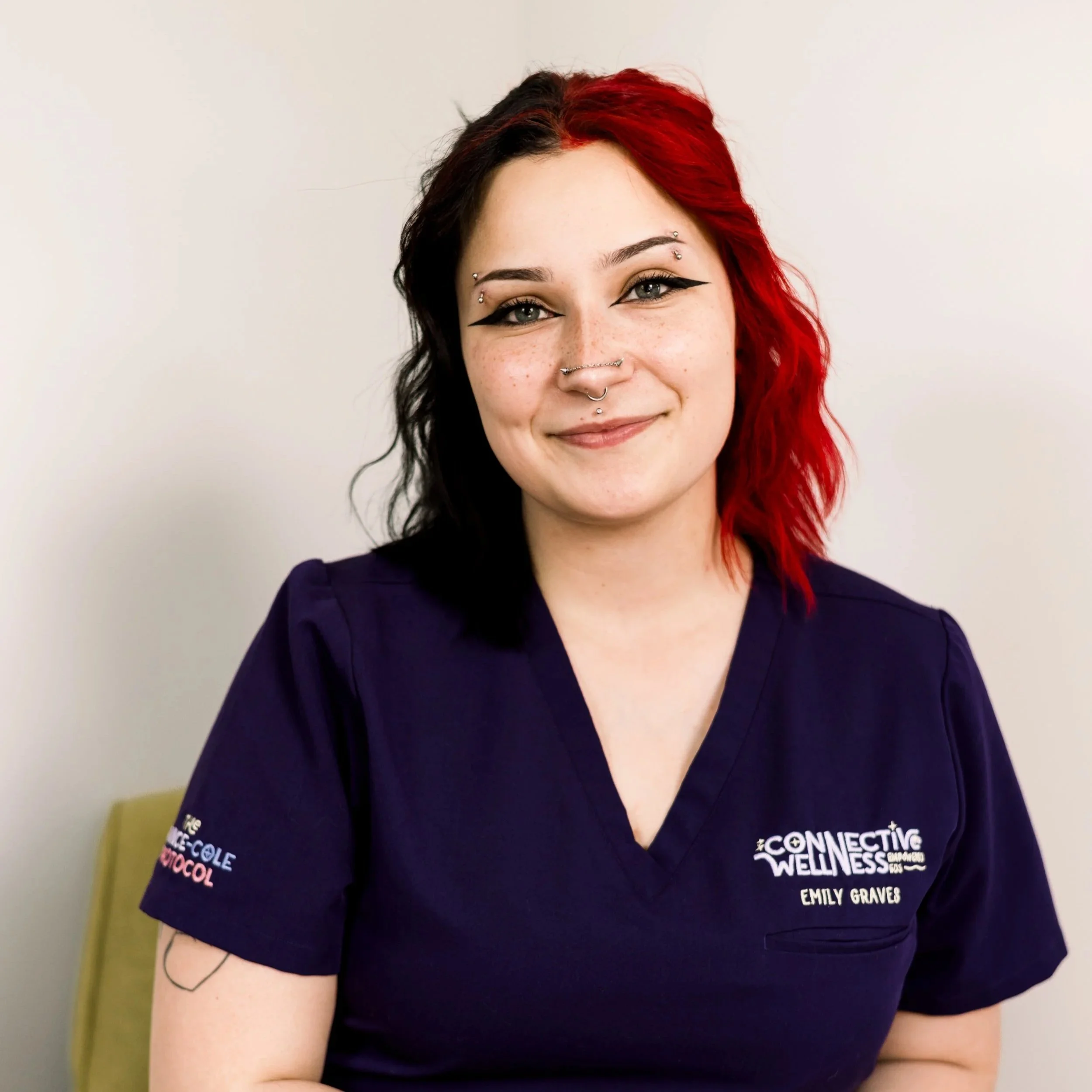A young woman with black and red hair and freckles, smiling, wearing a black tank top and gray cardigan, standing against a white background.