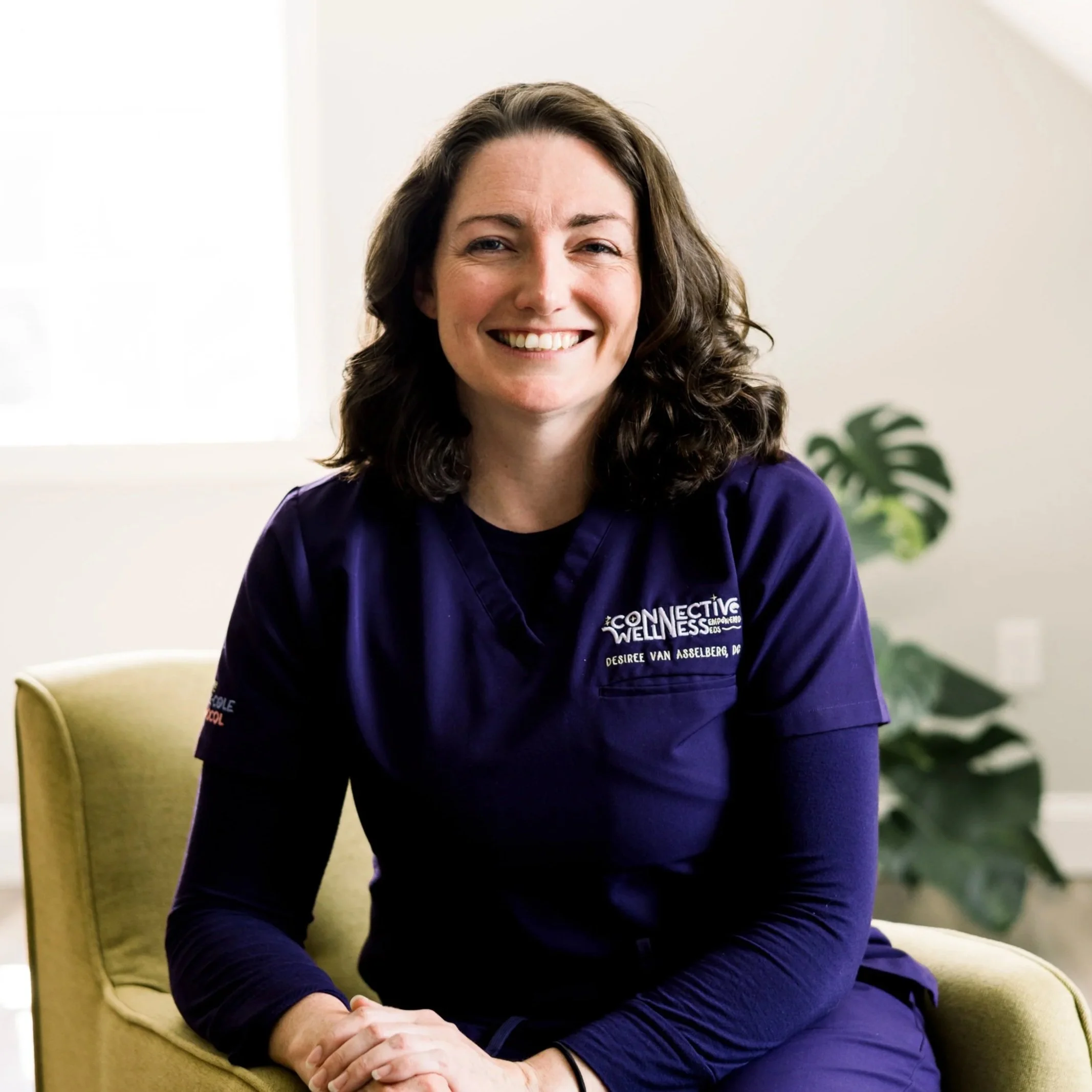 A woman with brown hair smiling, wearing a purple sweater, standing in front of a white wall.