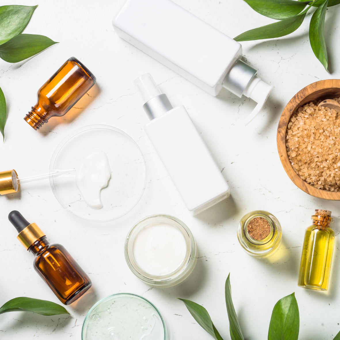Collection of skincare and cosmetic products, including bottles, jars, and dropper bottles on a white surface with green leaves and a wooden bowl of salt.