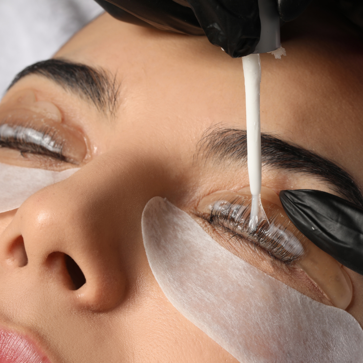 A person getting eyelash extensions applied with a technician using a dot of adhesive piped onto natural lashes, while wearing black gloves and a protective patch under the eye.