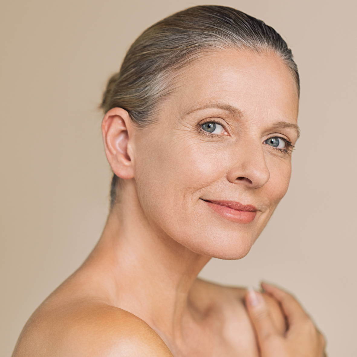 Close-up portrait of a middle-aged woman with short, slicked-back gray hair, blue eyes, and a soft smile, against a plain beige background.