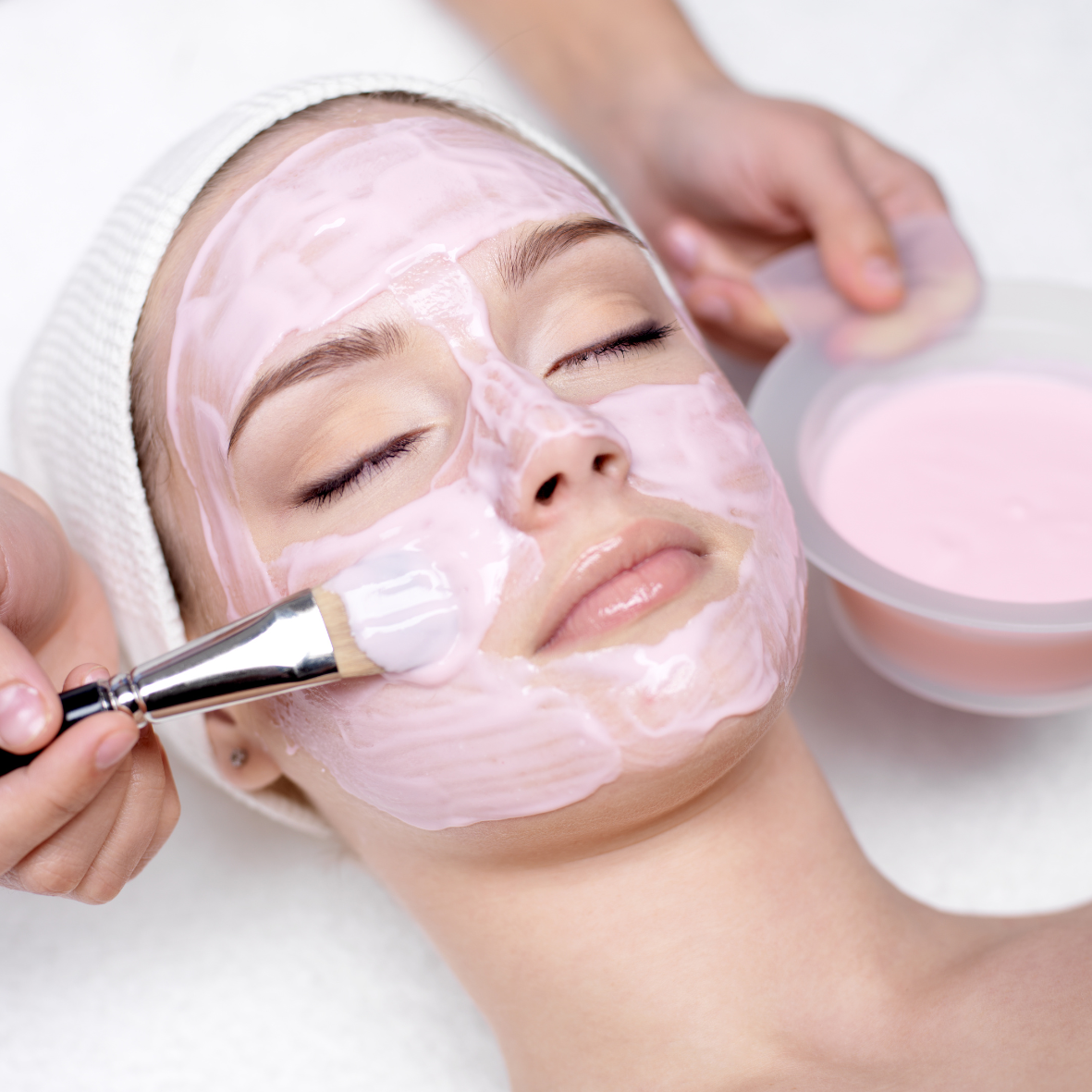 A woman is receiving a facial treatment, with her eyes closed and a pink facial mask being applied by a skincare professional using a brush. The woman is lying down with a towel wrapped around her head, and there is a bowl of the facial mask nearby.