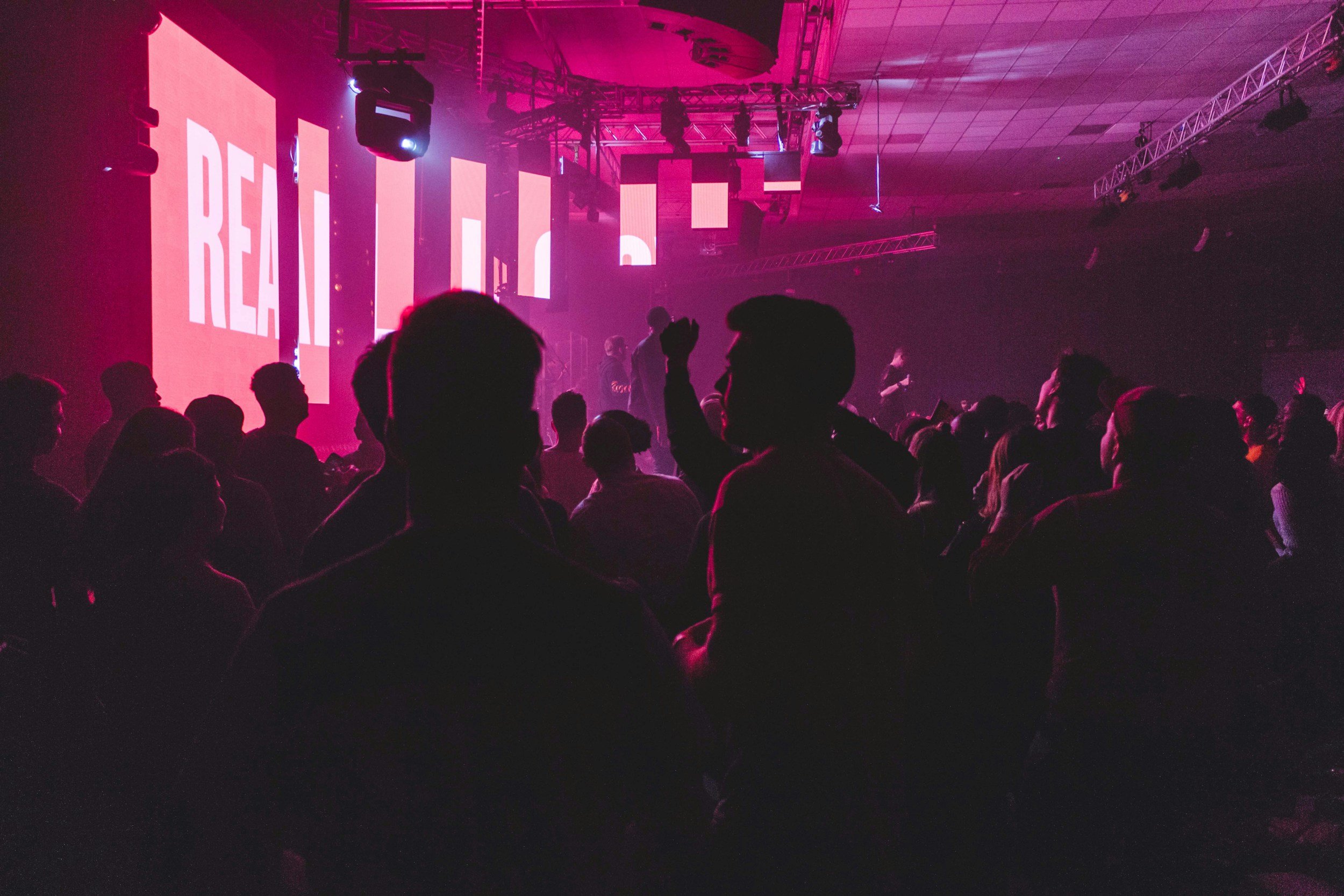 Crowd of people in a dark venue with pink and purple lighting, facing a stage with large screens displaying the word 'REAL' in bold white letters.