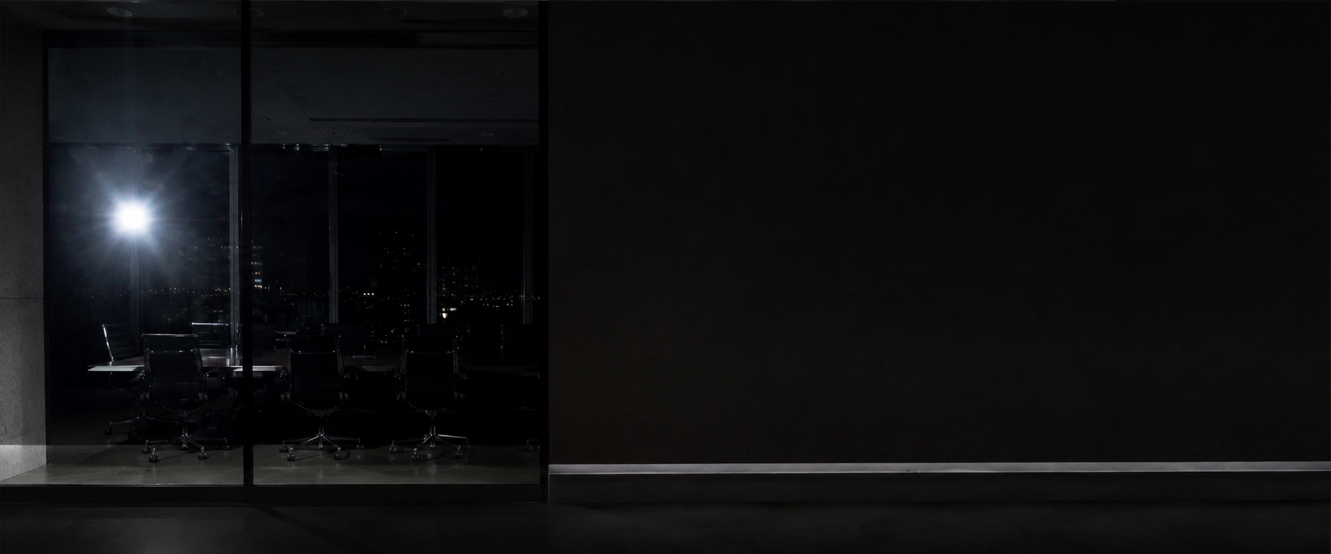 Dark conference room with glass walls, empty chairs, and city lights visible through the windows at night.