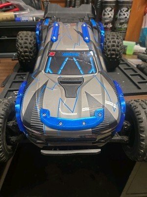 A remote-controlled car with a sleek blue and gray design, large wheels, and LED lights on the top, placed on a black surface in a room with various objects in the background.