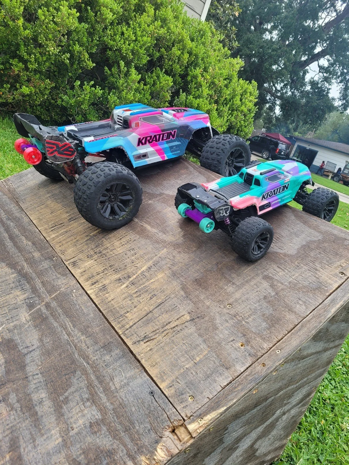 Two colorful remote-controlled off-road trucks with large tires are on a wooden surface outdoors, with green bushes and a house in the background.