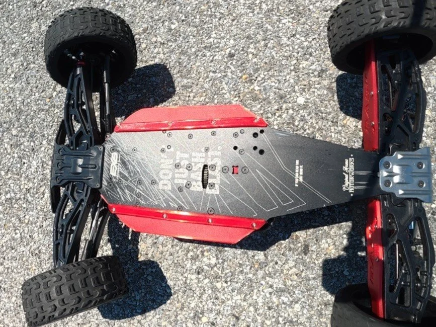 A red and black remote-controlled car with exposed chassis and large off-road tires on a gravel surface.