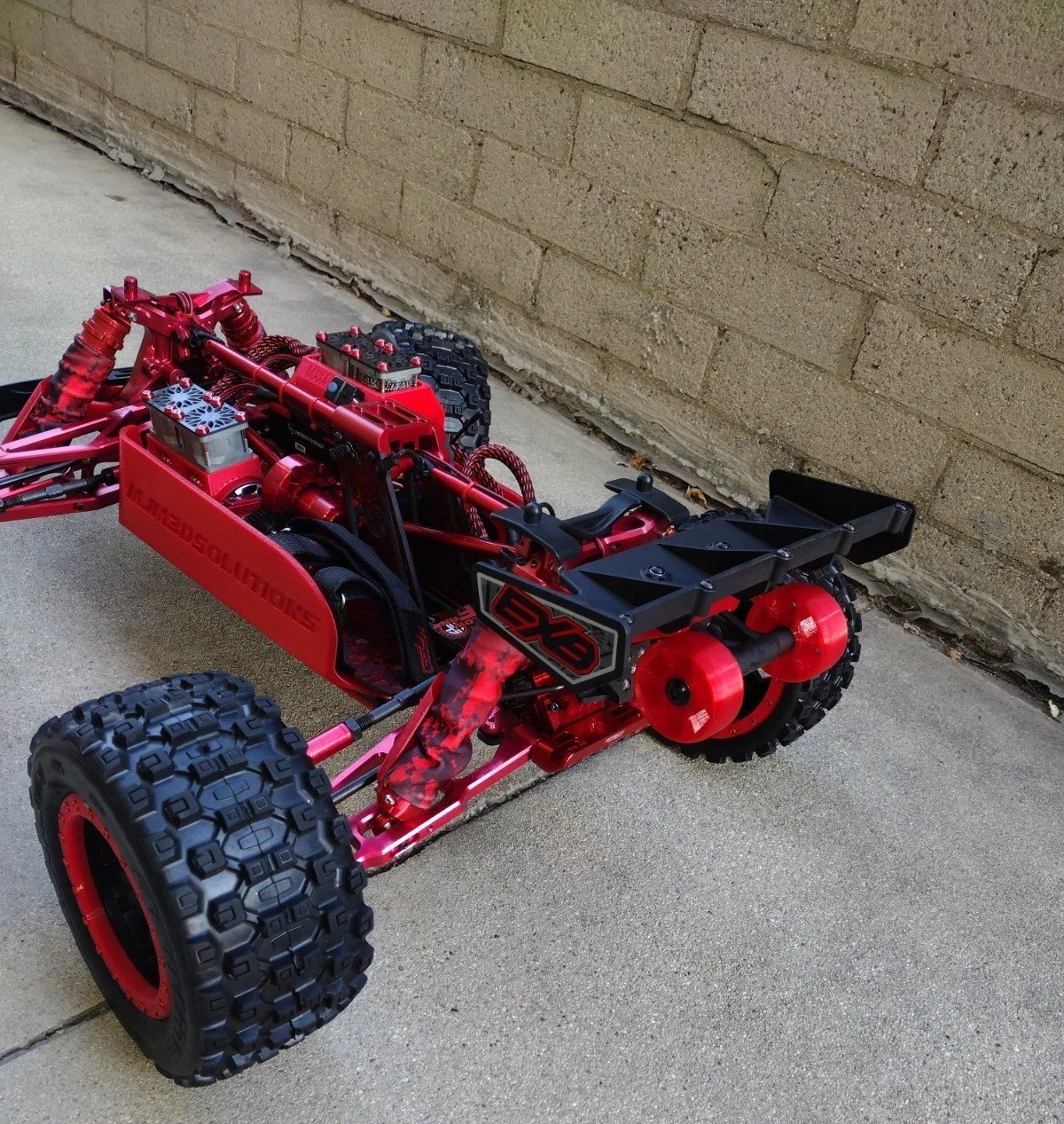 Red RC off-road vehicle with large tires, black and red suspension, and various components, parked on concrete next to a brick wall.