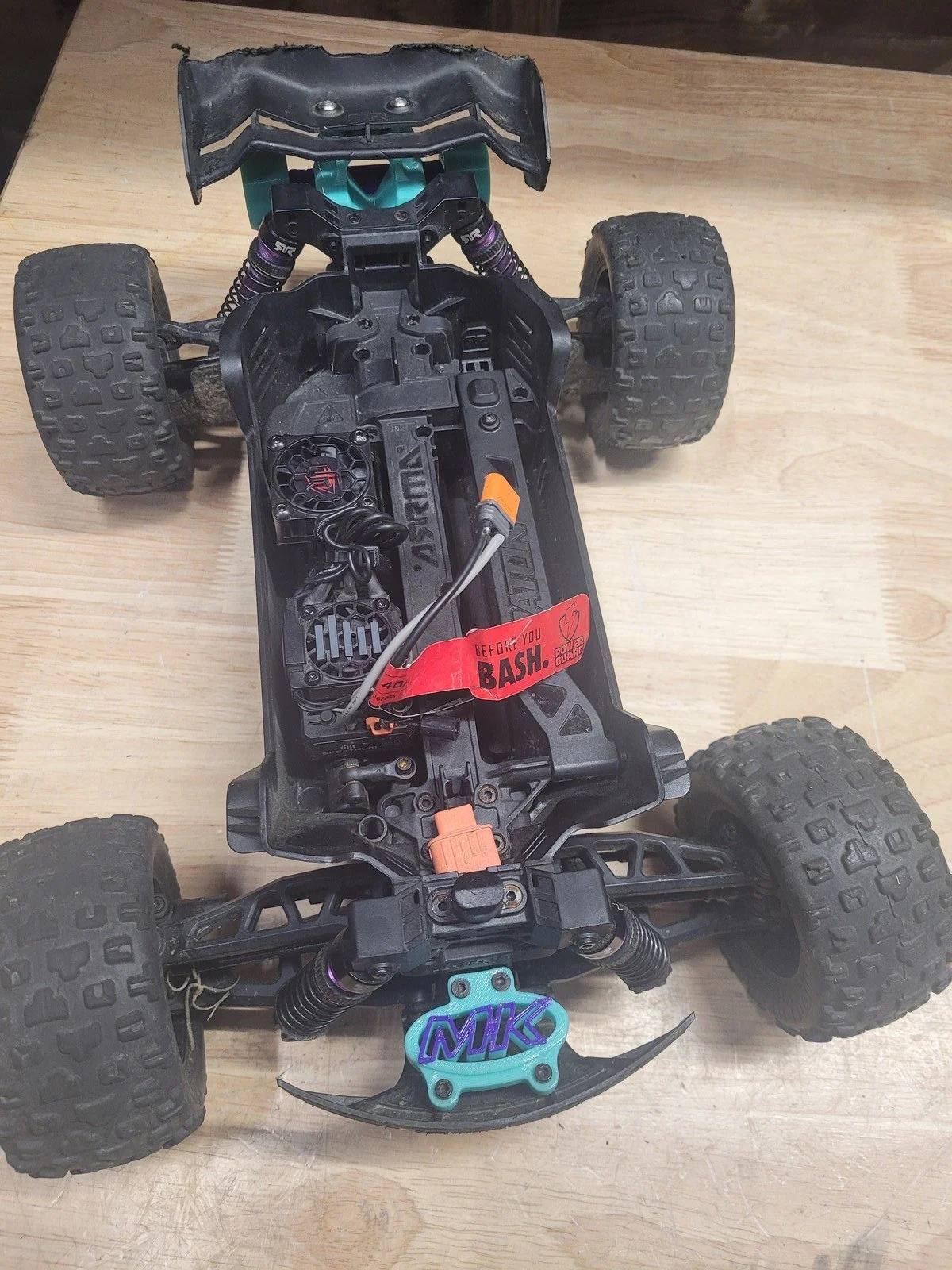 The image shows the underside of a remote-controlled off-road vehicle with four large rugged tires, a partially exposed chassis, and wiring. The vehicle features a red warning label that says 'Before You Bash.'
