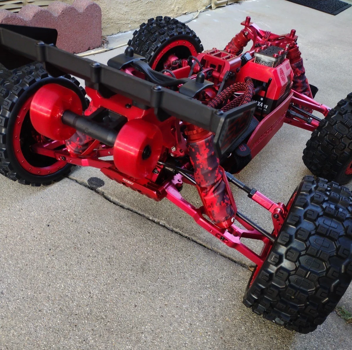 Red RC off-road truck chassis with large tires, visible electronic components, and suspension system on a concrete surface.