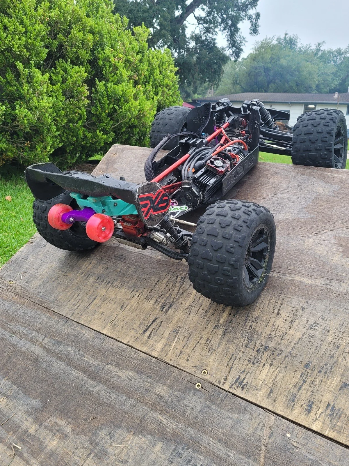 A remote-controlled off-road car with large tires, a black chassis, and visible electronic components, resting on a wooden surface outdoors with green bushes and trees in the background.