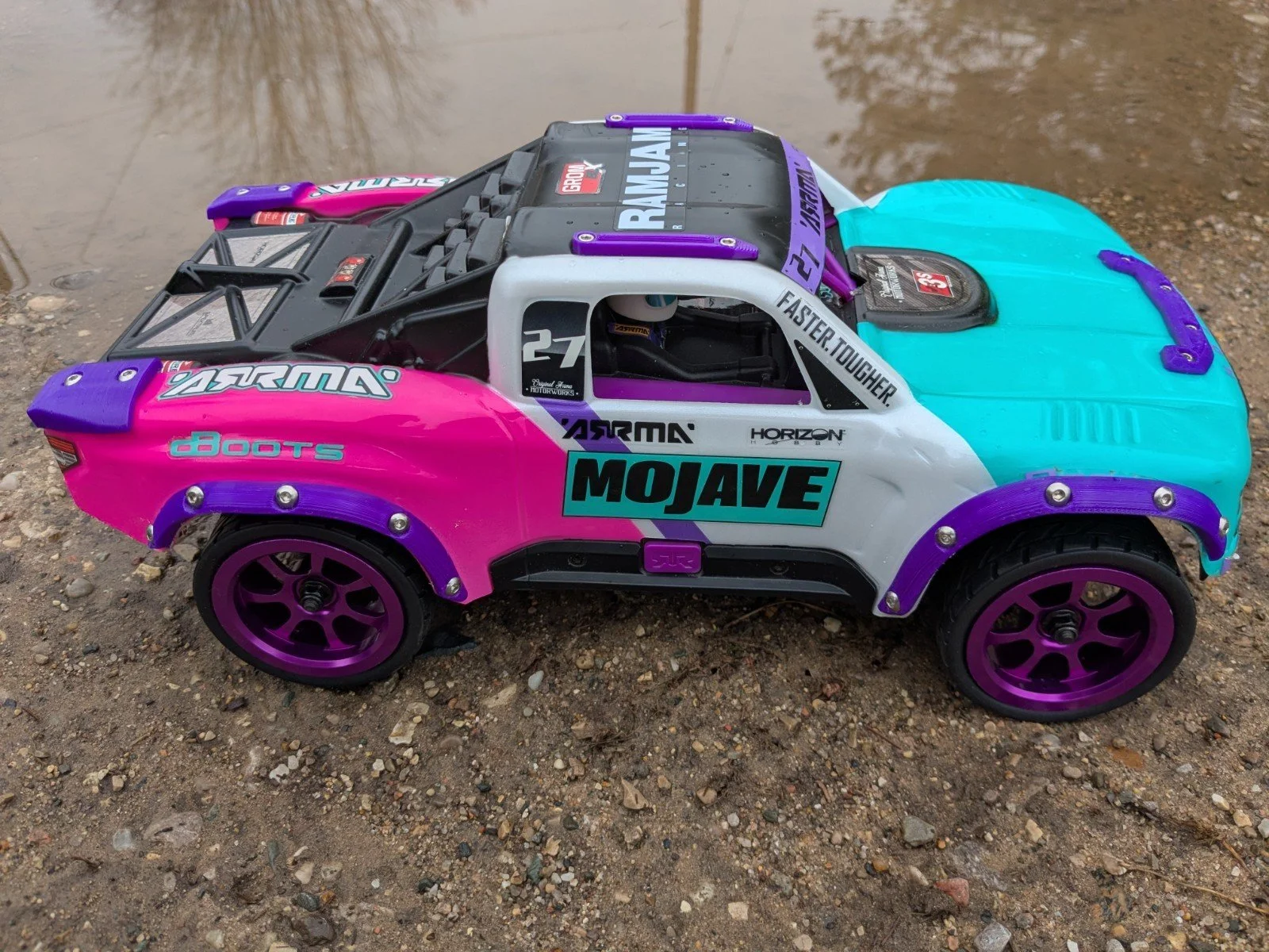 Colorful remote-controlled car with pink, turquoise, and purple accents on gravel near water.