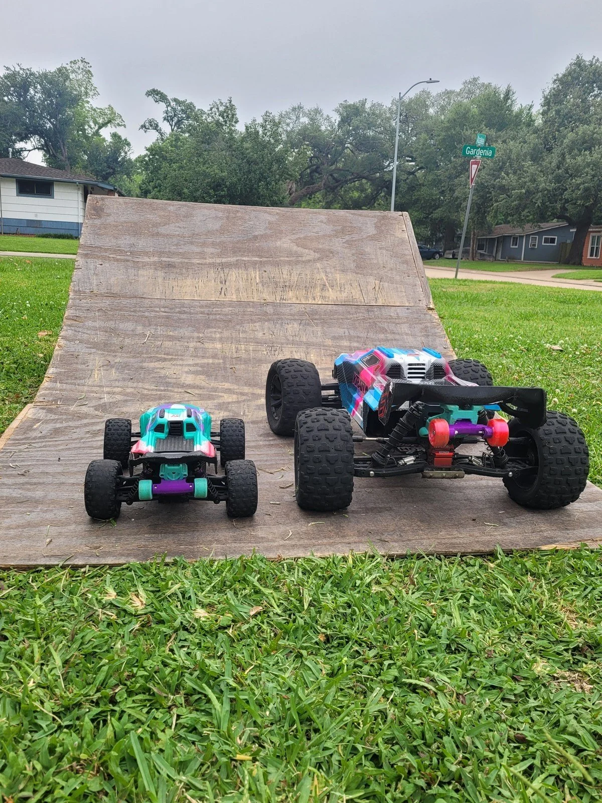 Two RC cars, one small and one large, on a wooden ramp in a grassy area with trees and houses in the background.