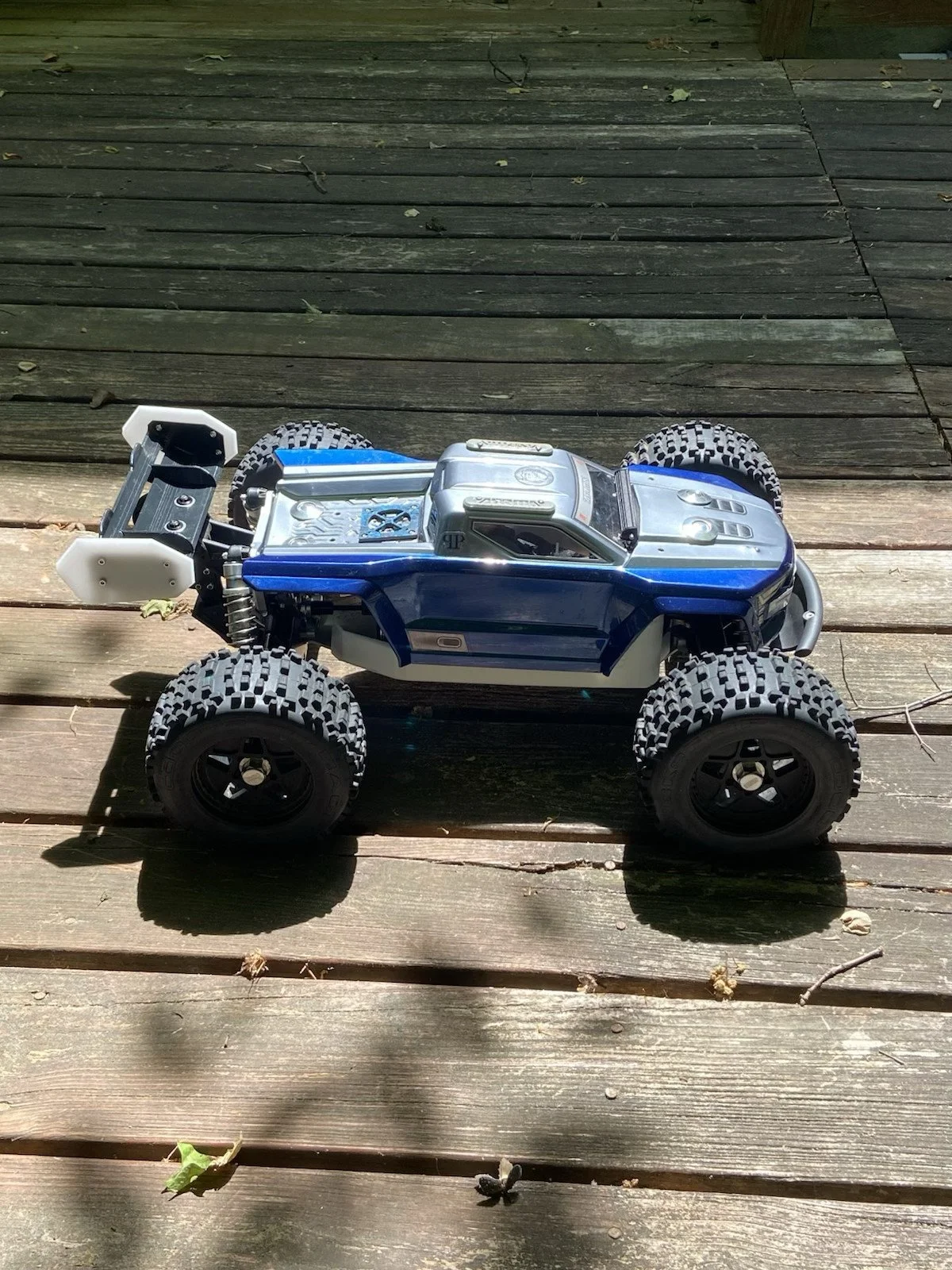 A remote-controlled toy off-road truck with large knobby tires and a blue and silver body on a wooden deck.