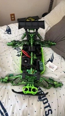 Green and black remote-controlled helicopter with a camera mounted on top, placed on a bed with patterned sheets and pillows.