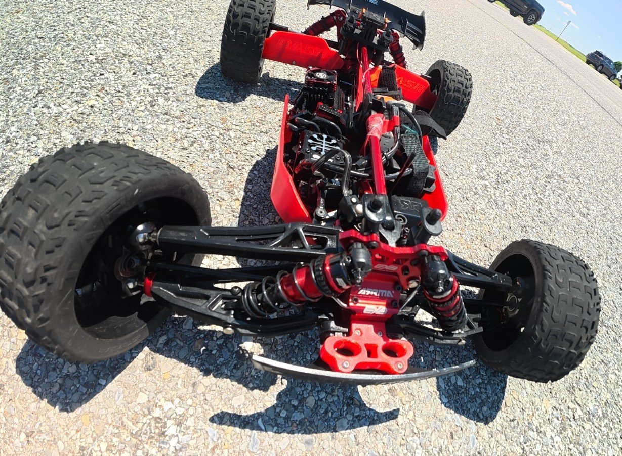 Red remote-controlled car with exposed chassis, suspension, and tires on a gravel surface.