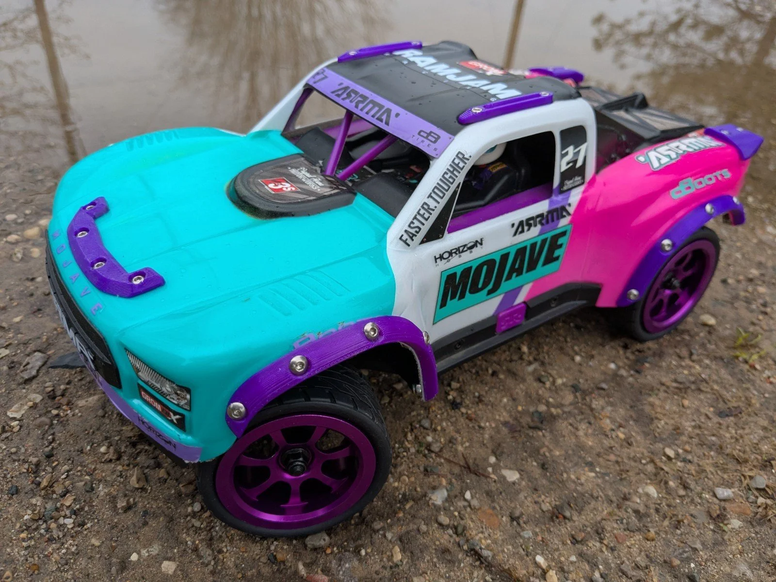 Colorful remote-controlled car with a turquoise, pink, white, and purple paint job, placed on gravel ground near water.