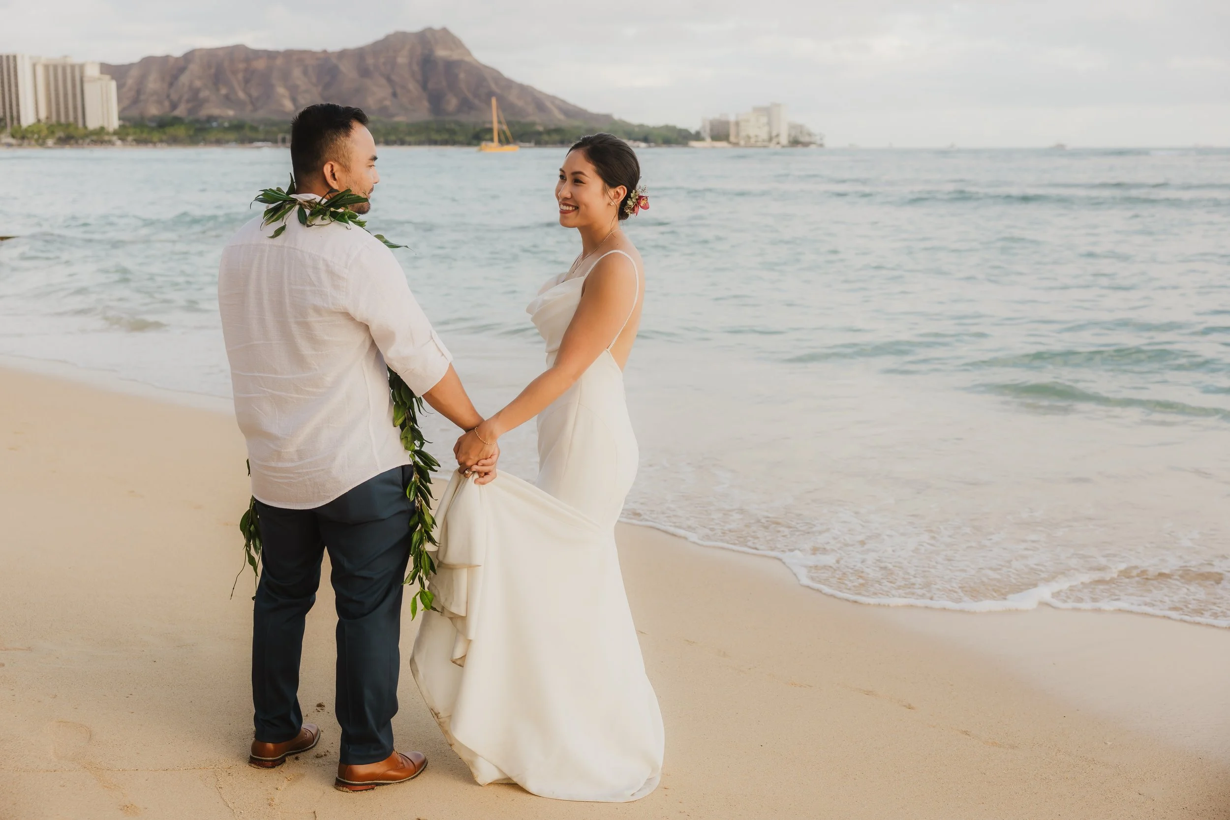 A couple gets married on a beach, holding hands and smiling at each other with the ocean and mountains in the background.