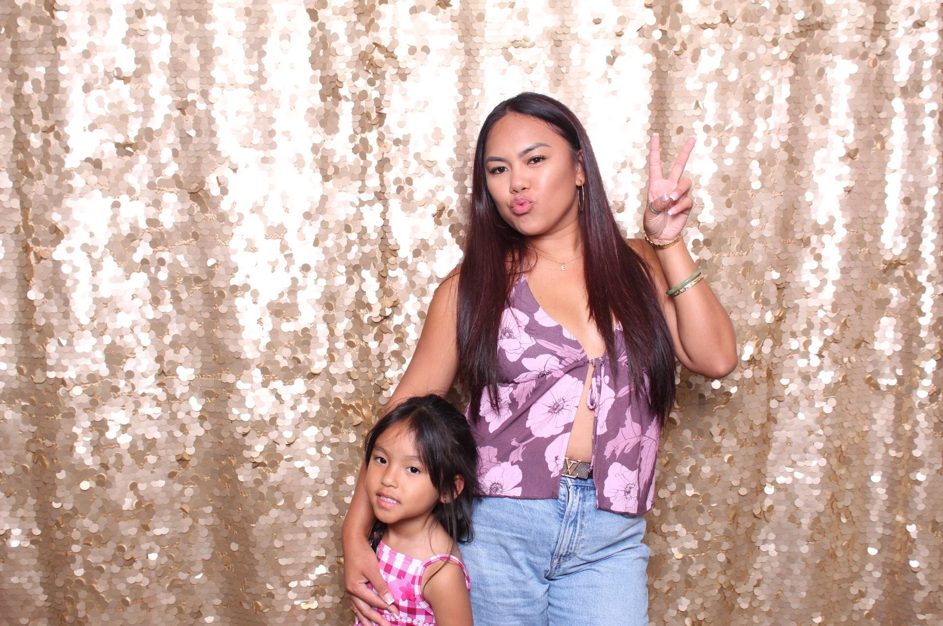 Two young girls and a woman standing in front of a gold sequin backdrop. The woman is making a peace sign and puckering her lips, wearing a purple floral top and jeans. One girl is in a pink checkered dress, and the other girl is partially visible at the bottom, wearing a pink checkered top.