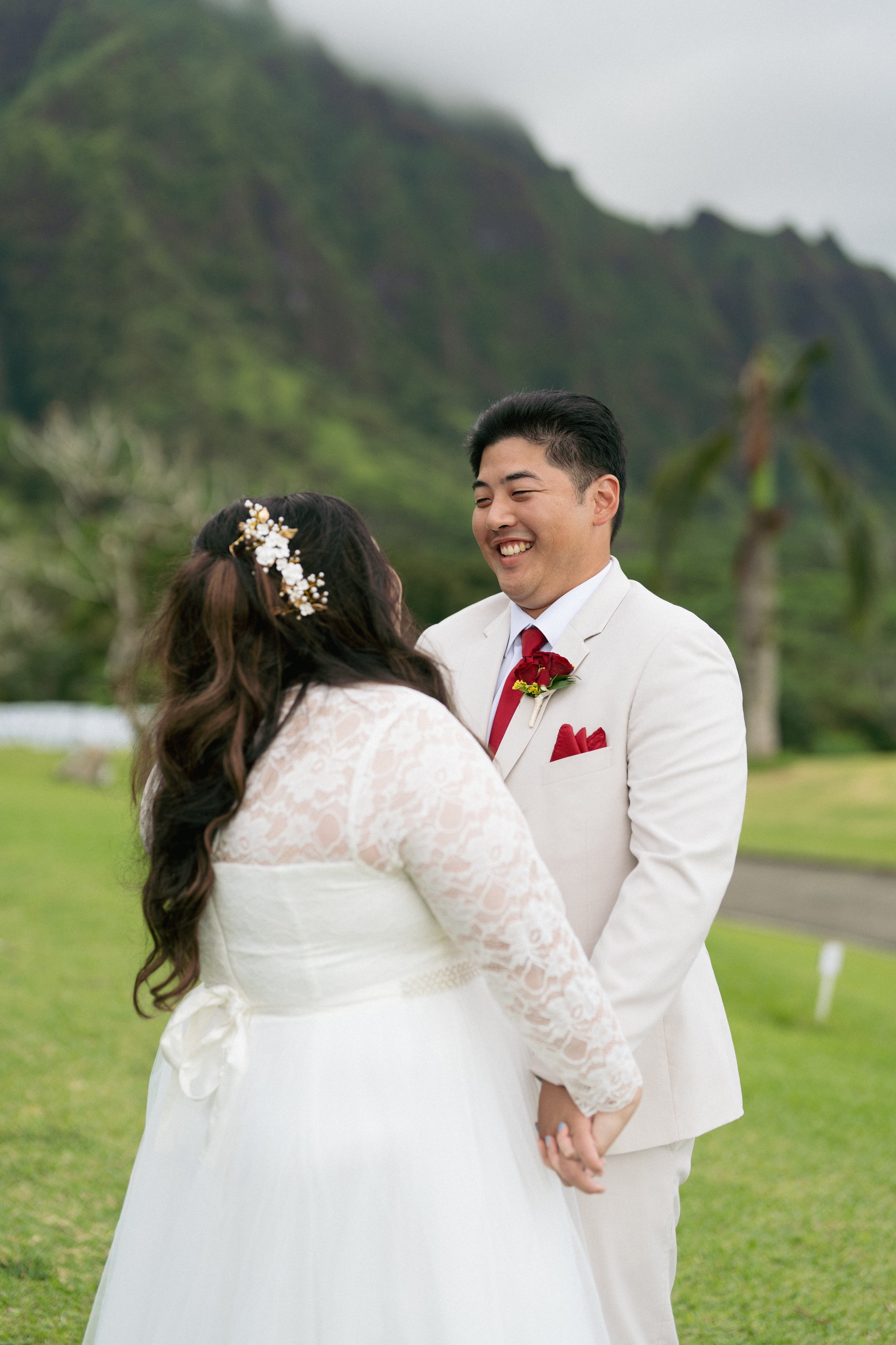 A couple holding hands and smiling at each other outdoors during a wedding, with a green mountain and cloudy sky in the background. Justyn Tabaniag oahu hawaii