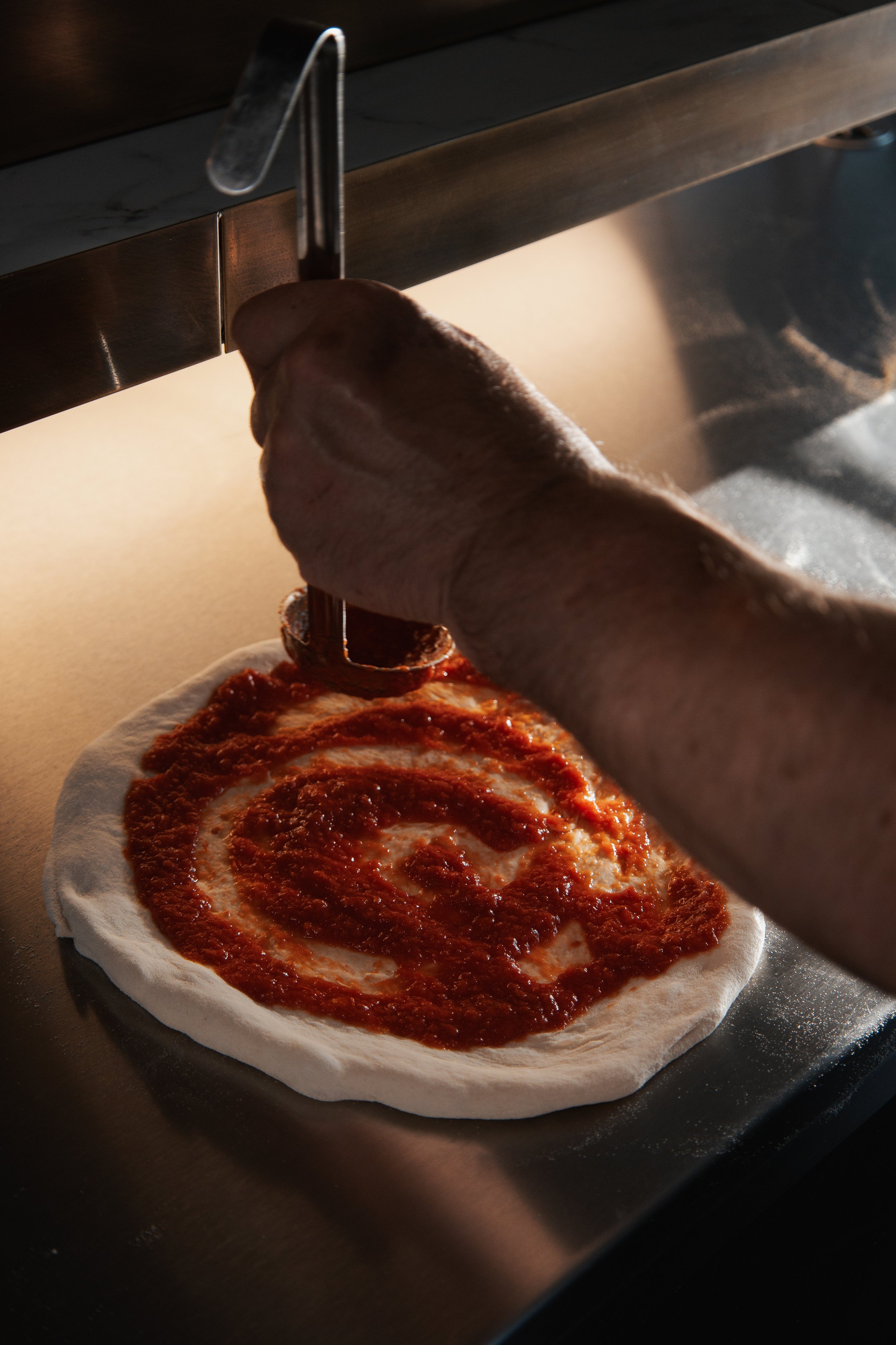 A person is spreading tomato sauce on pizza dough using a ladle in a kitchen.