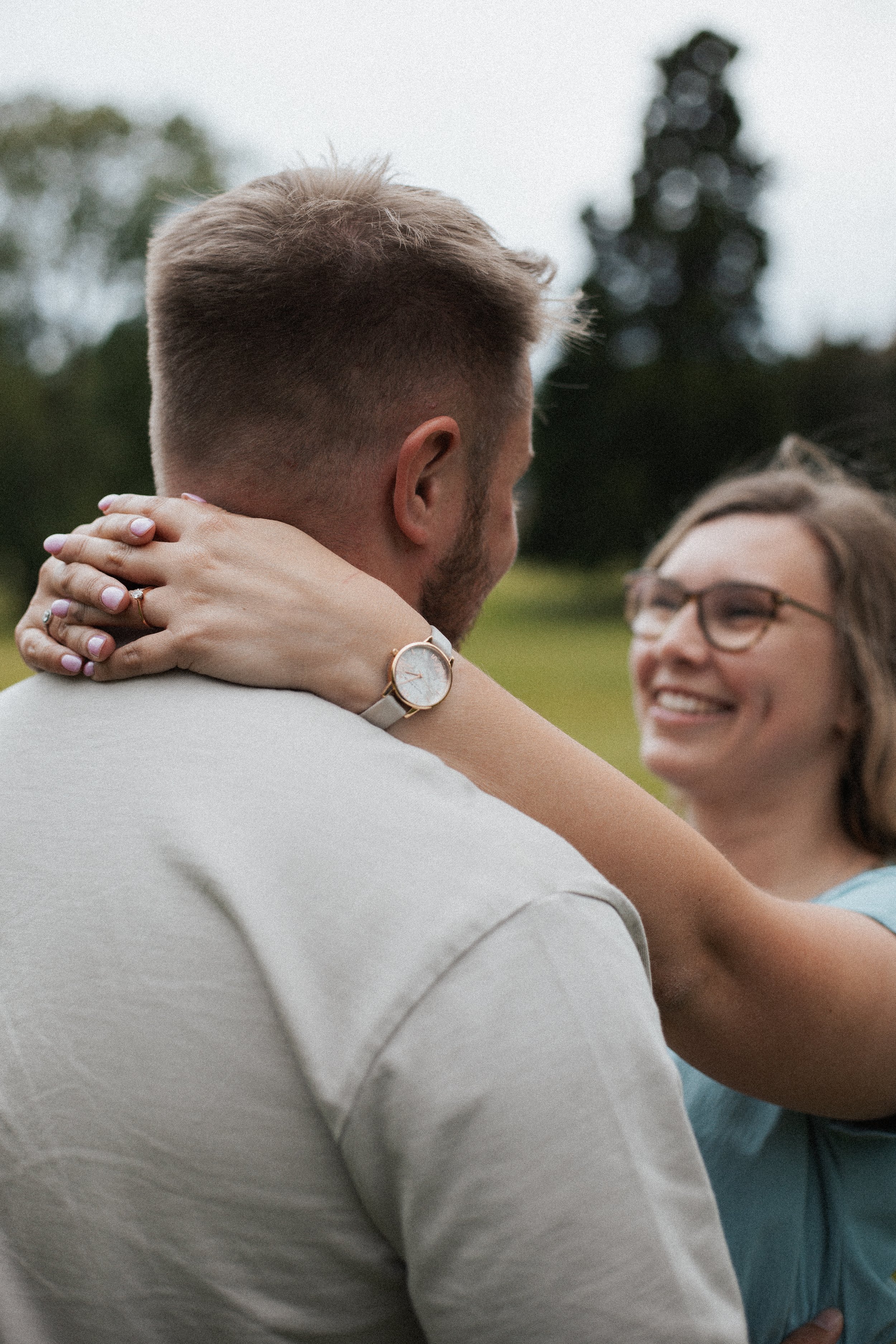 A couple is embracing outdoors on a cloudy day, with the woman smiling and wearing glasses, while the man is turned away from the camera. She has her arms around his neck, showing a watch on her wrist.