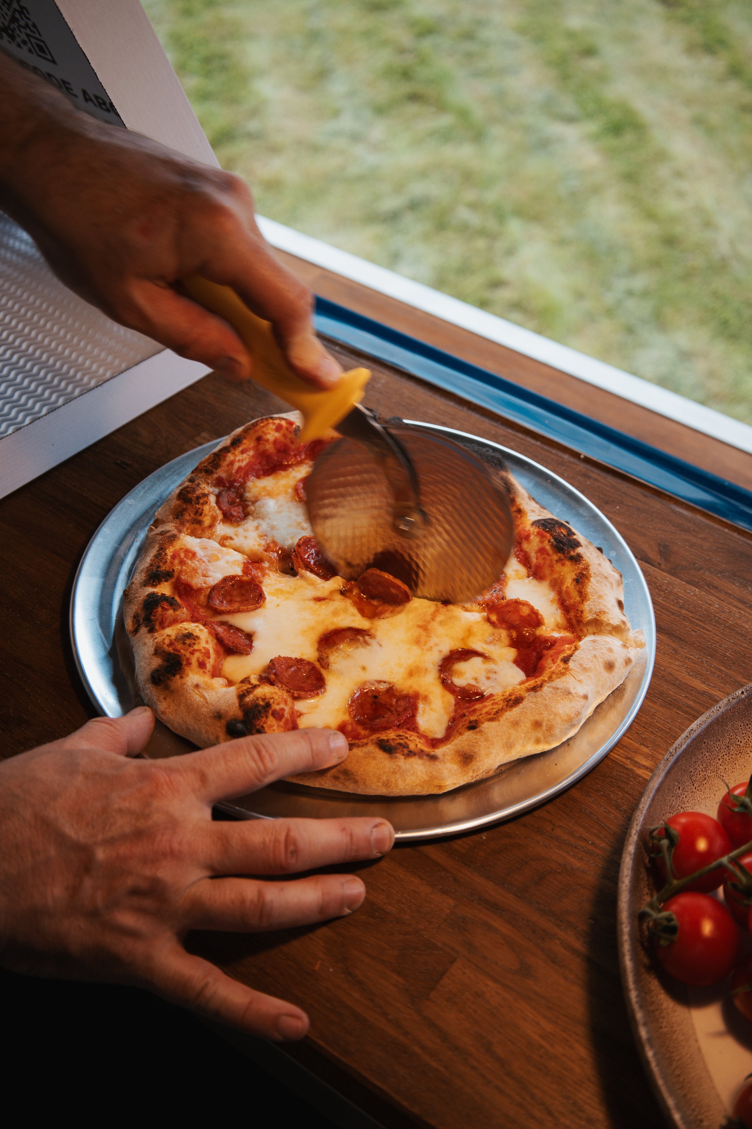 Hands slicing a pepperoni pizza with a pizza cutter on a round metal tray, on a wooden table near a window with a grassy yard outside.