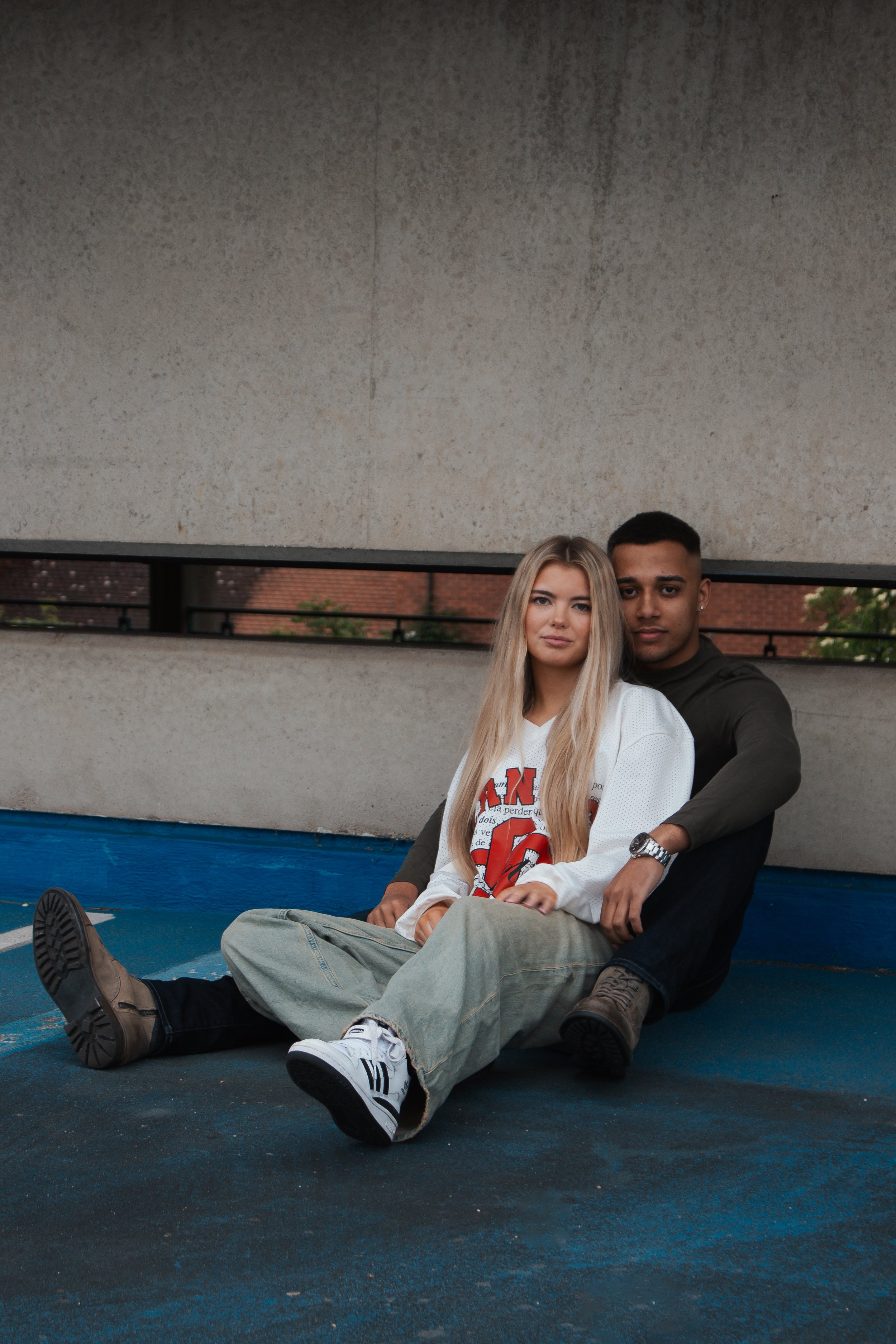 A young couple sitting on the ground in an urban setting, with a girl in a white sports jersey and a boy in dark clothing, posing for a photo.