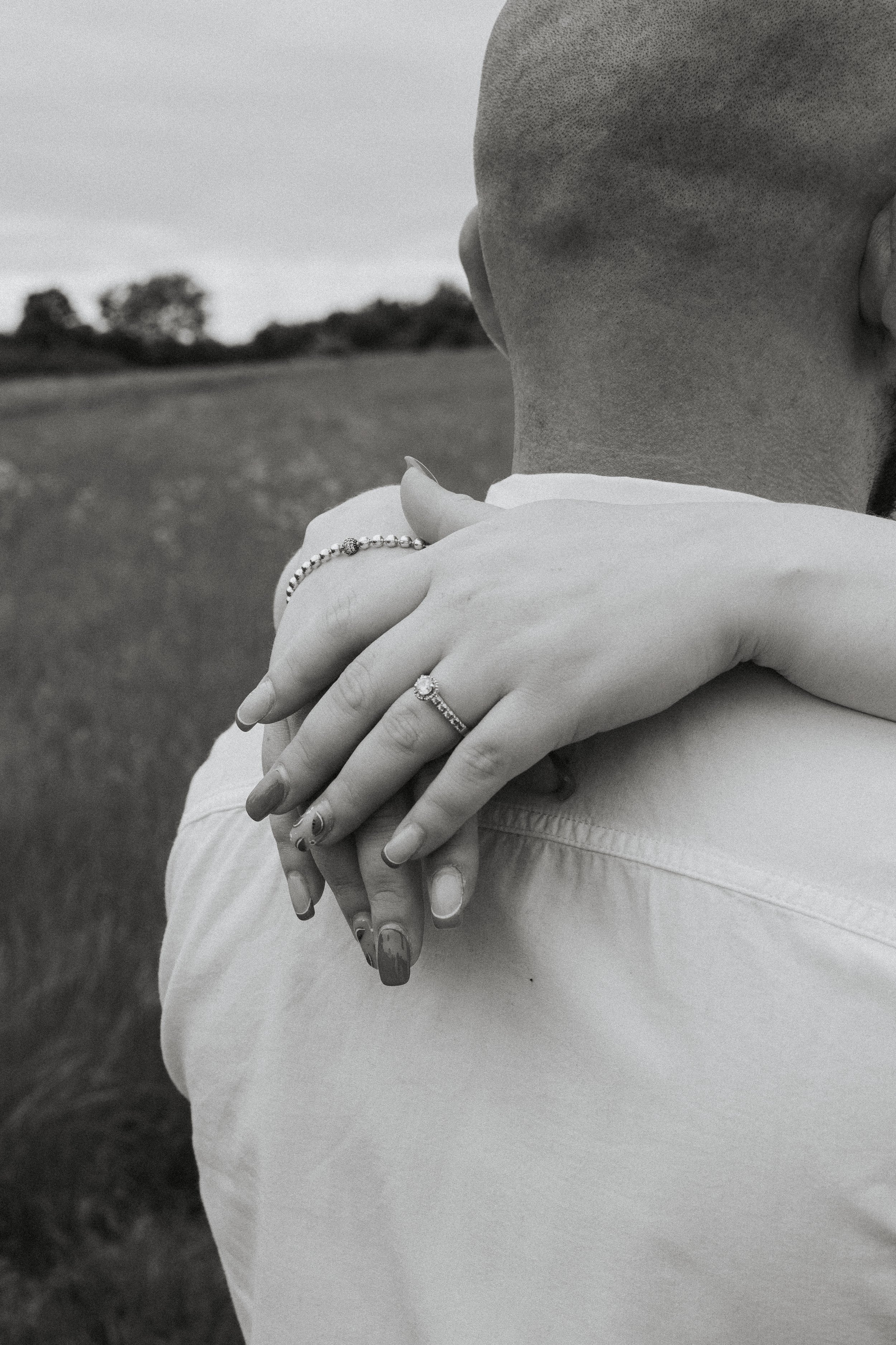 Close-up of a couple embracing outdoors, showing their hands with wedding rings, in black-and-white. The man's head and shoulders are visible from behind, while the woman's hands are resting on his shoulder.