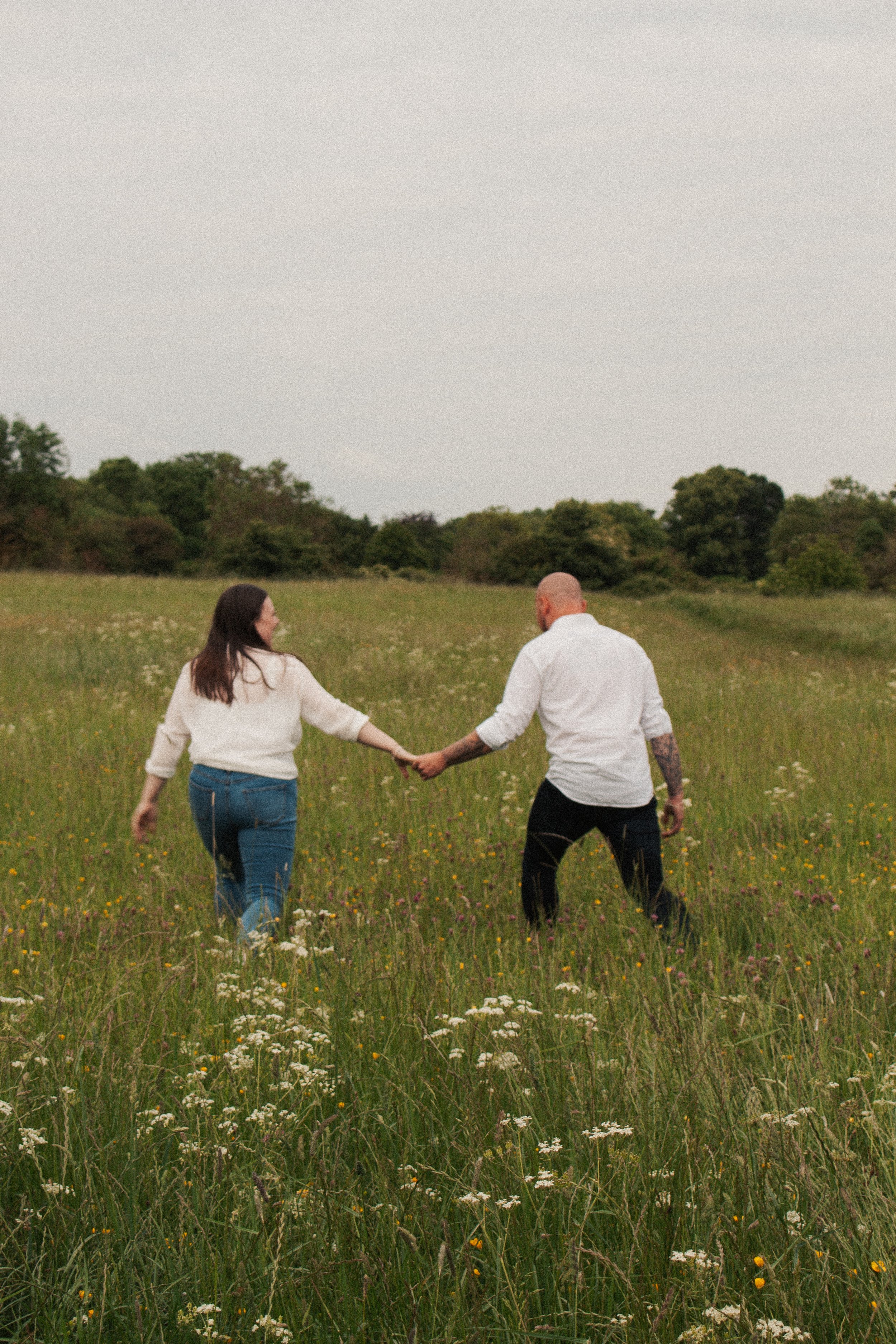 A couple holding hands and walking through a field of wildflowers on a cloudy day.