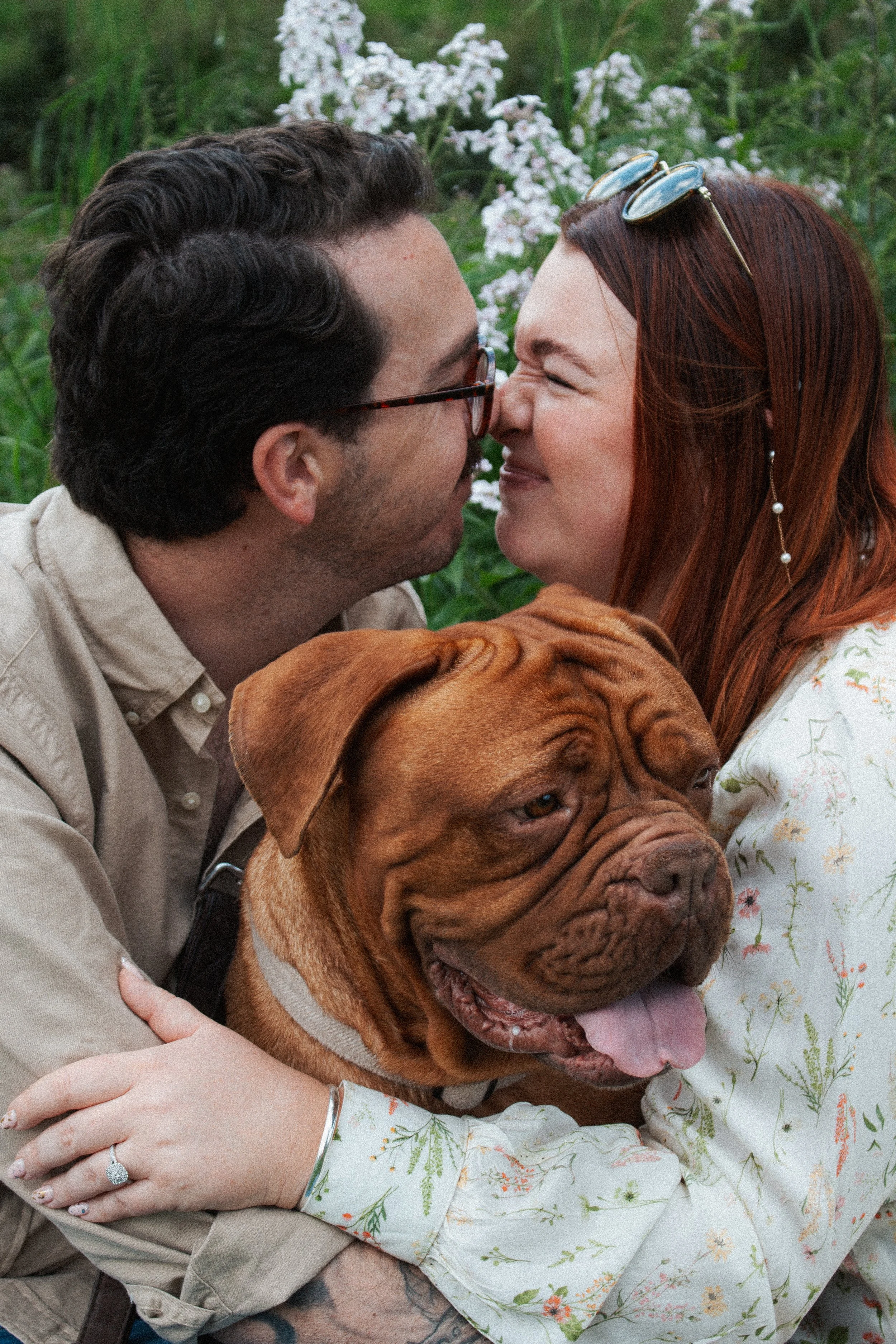 A couple is sharing a close moment outdoors, nose-to-nose, with a large brown dog between them. The woman has red hair and wears glasses on her head, and the man has dark hair and glasses. The dog looks content with its tongue partially out.